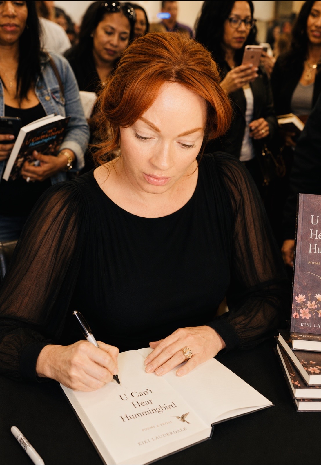 Red-haired woman signing a book at a table, surrounded by people, some holding books and phones.