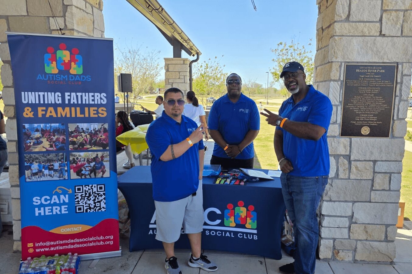 Three men in blue shirts stand behind a table with a colorful logo, outdoors near a stone wall and a banner, smiling and posing. Word count: 20