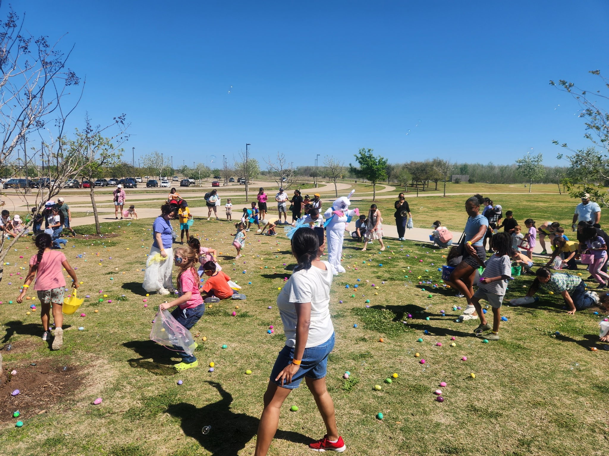 Children and adults gather outdoors on grass, participating in an egg hunt on a sunny day with trees and a blue sky.