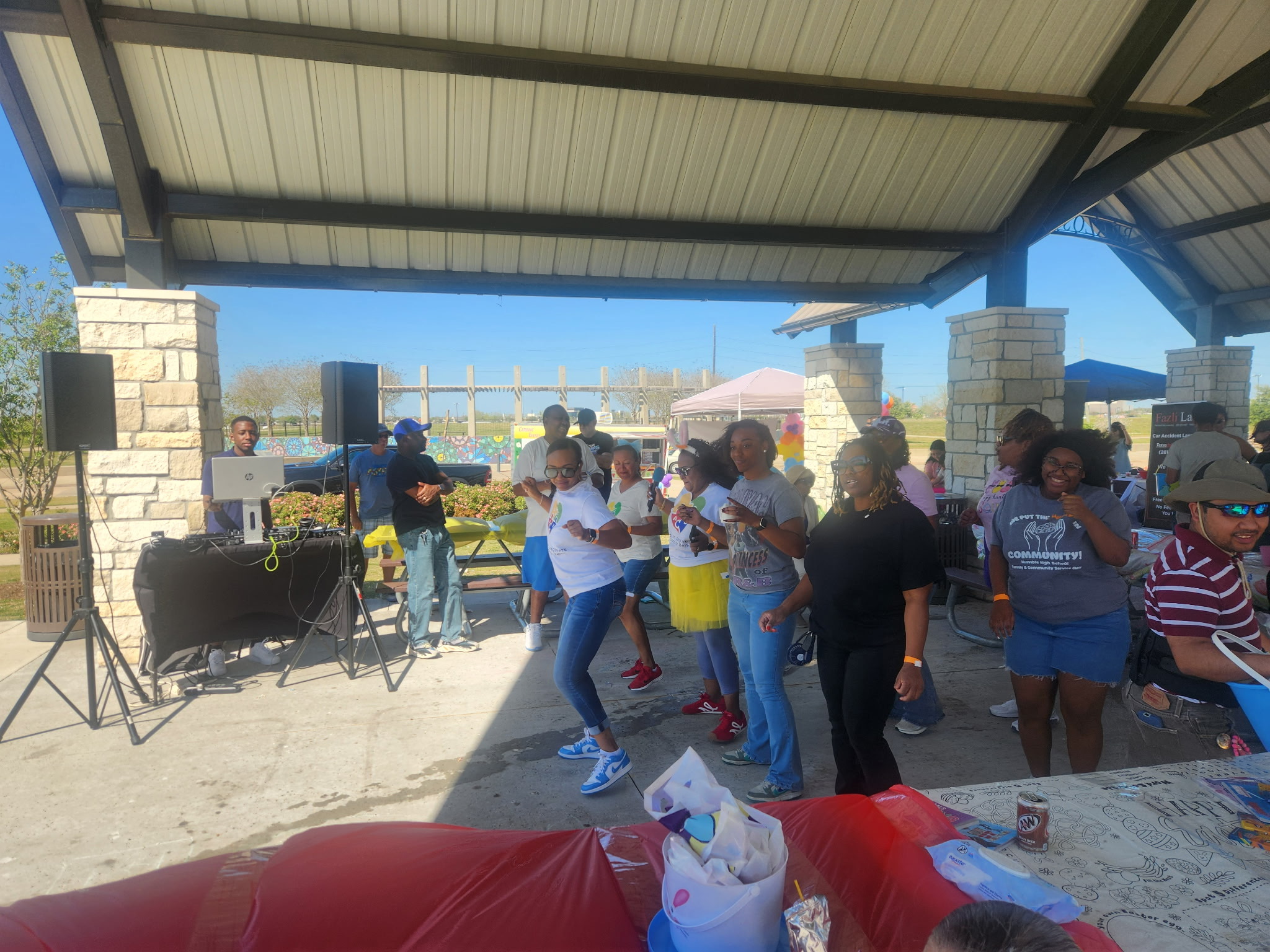 Group of people gathered outdoors under a pavilion, some dancing, with speakers and tables nearby, sunny day.