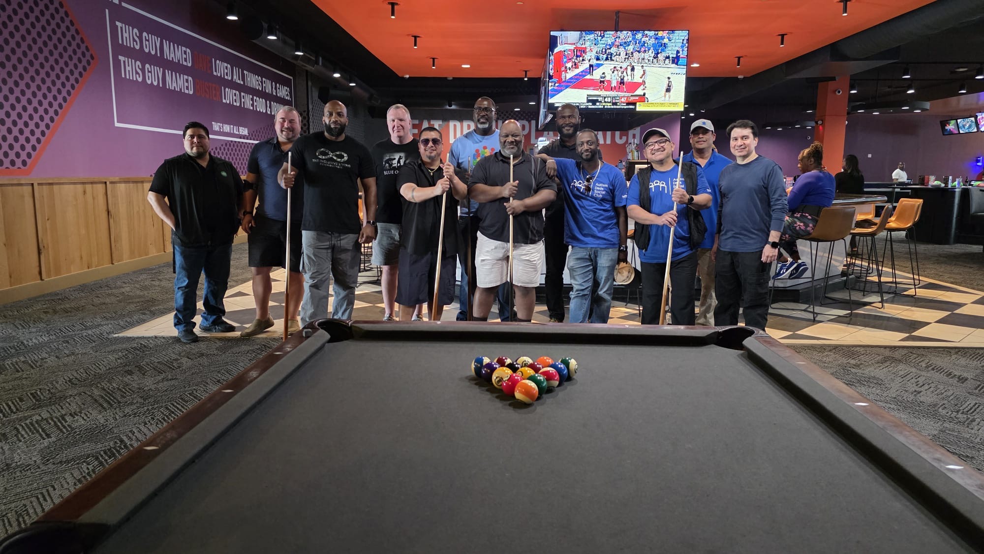 Group of people standing around a pool table in a gaming area, with a TV screen in the background.