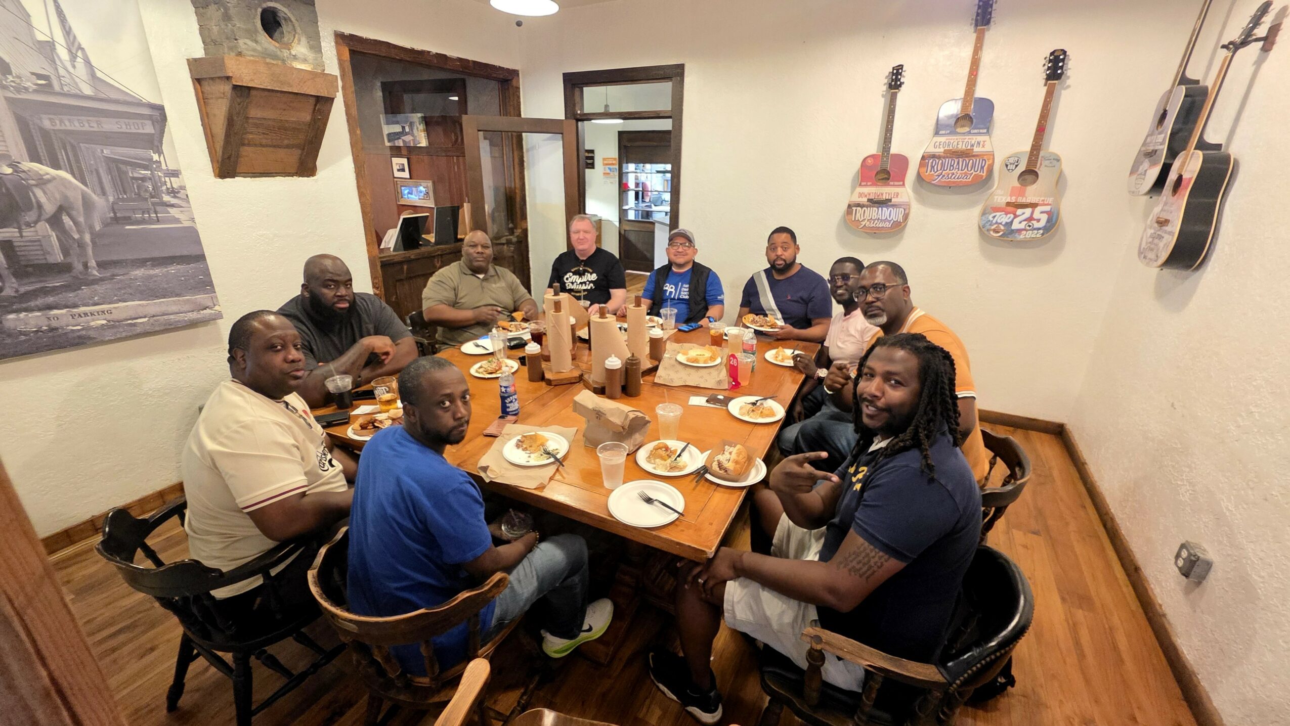 Group of people sitting around a dining table in a restaurant, eating and talking, with guitars hanging on the wall.