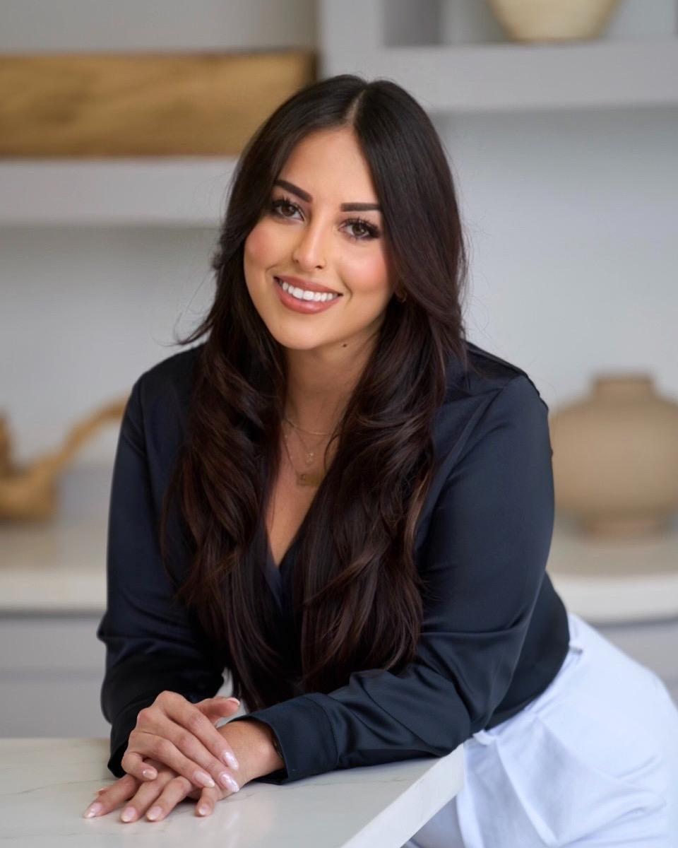 Young woman with long dark hair smiling, leaning on a white surface in a modern kitchen, wearing a dark top.