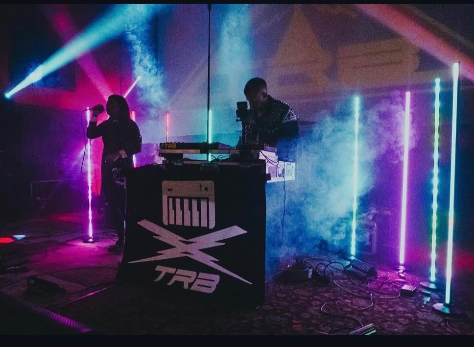 Two people perform on stage with colorful lights and fog, DJ equipment in front, dark background, vibrant atmosphere.