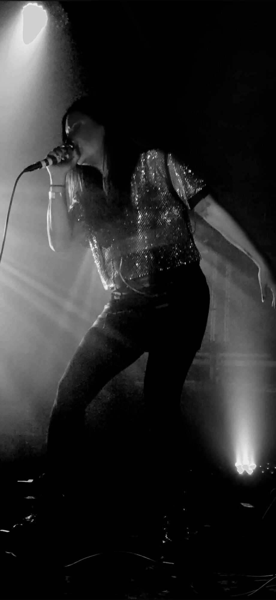 Person singing into a microphone on stage with dramatic lighting, wearing a sparkly top and dark pants.