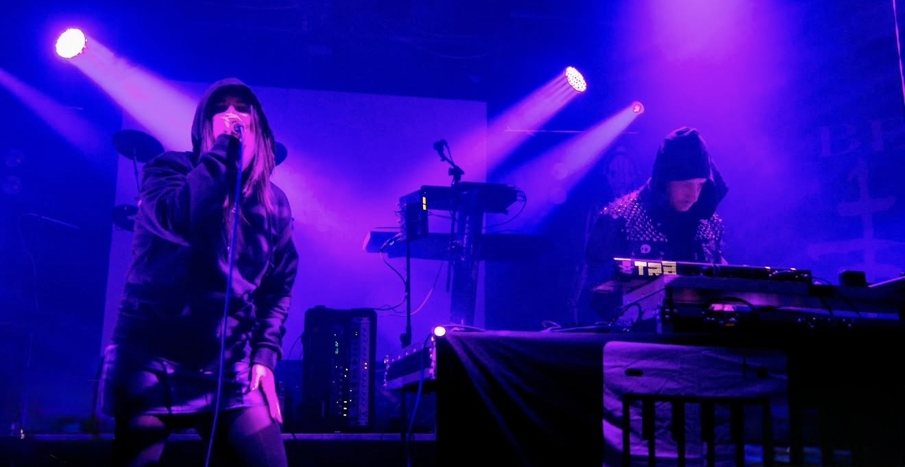 Two musicians perform on stage with purple and blue lighting, one singing into a microphone and the other playing equipment.