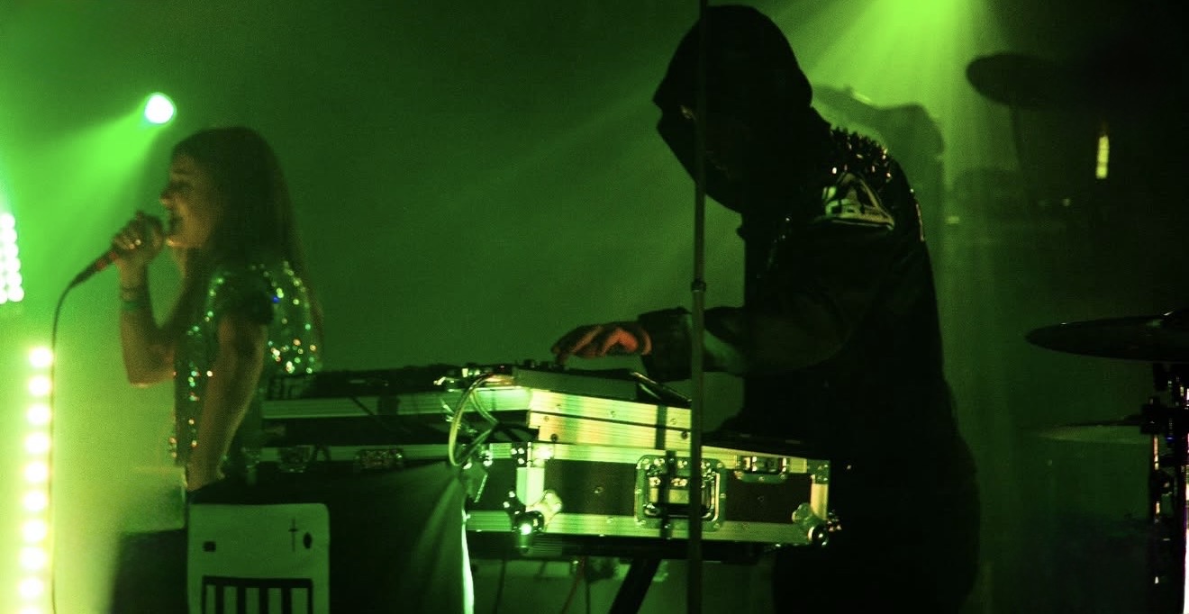 Two performers on stage with green lighting, one singing into a microphone and the other operating DJ equipment.