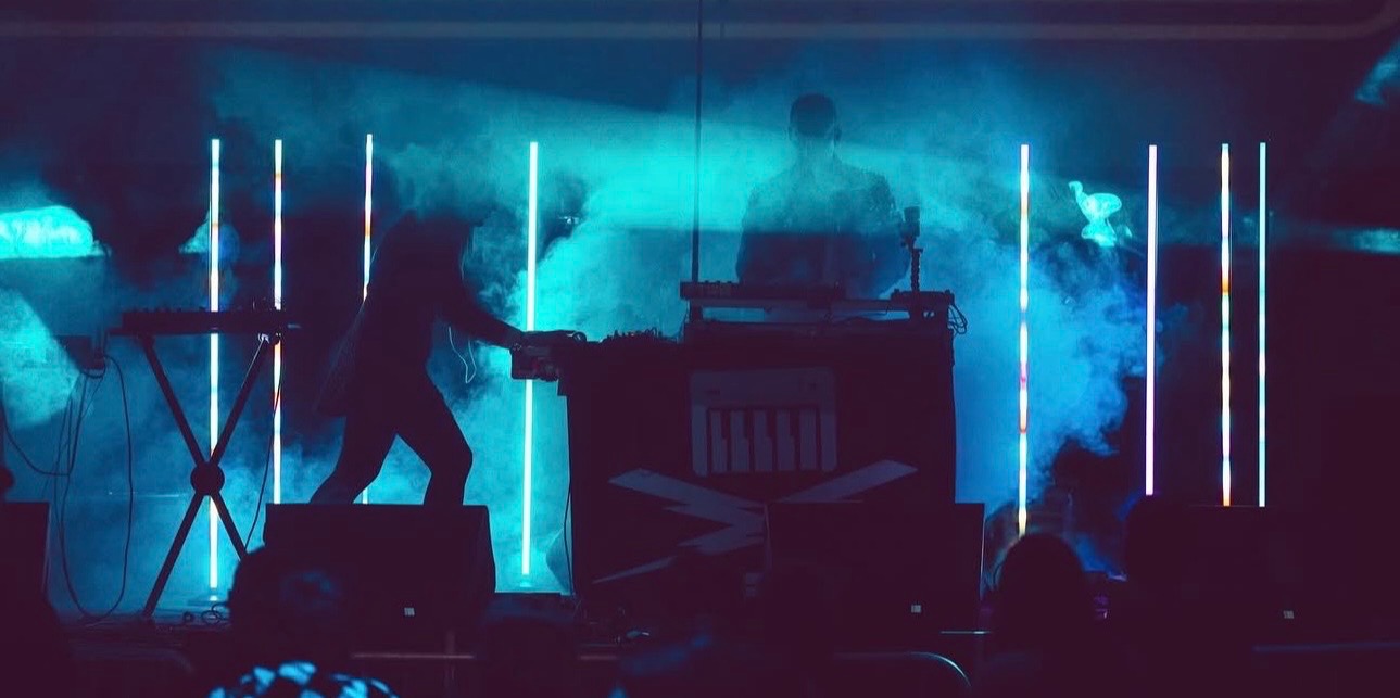 Two performers on stage with blue lighting and smoke effects, one reaching out to the other, electronic equipment in the background.
