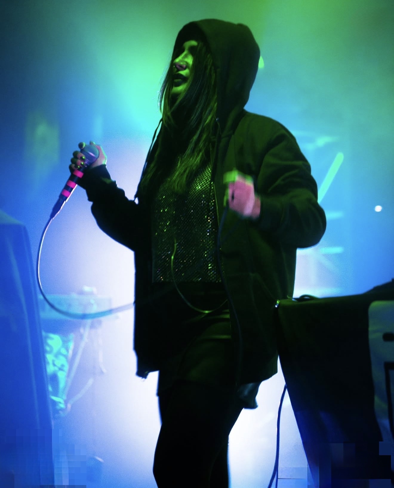Person with long hair wearing a hoodie and jacket, holding a microphone, illuminated by green and blue lights.