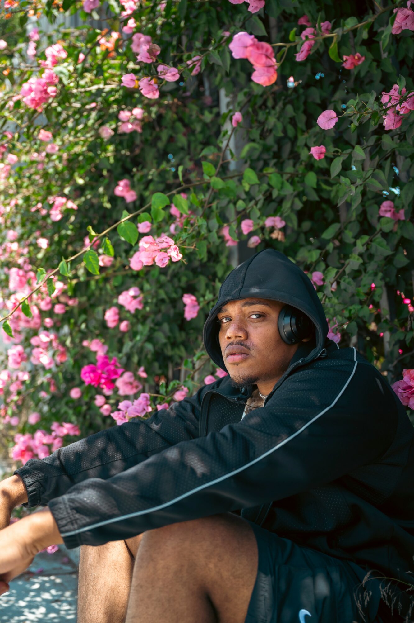 Young person wearing a black hoodie and headphones, sitting outdoors near pink flowering bushes.