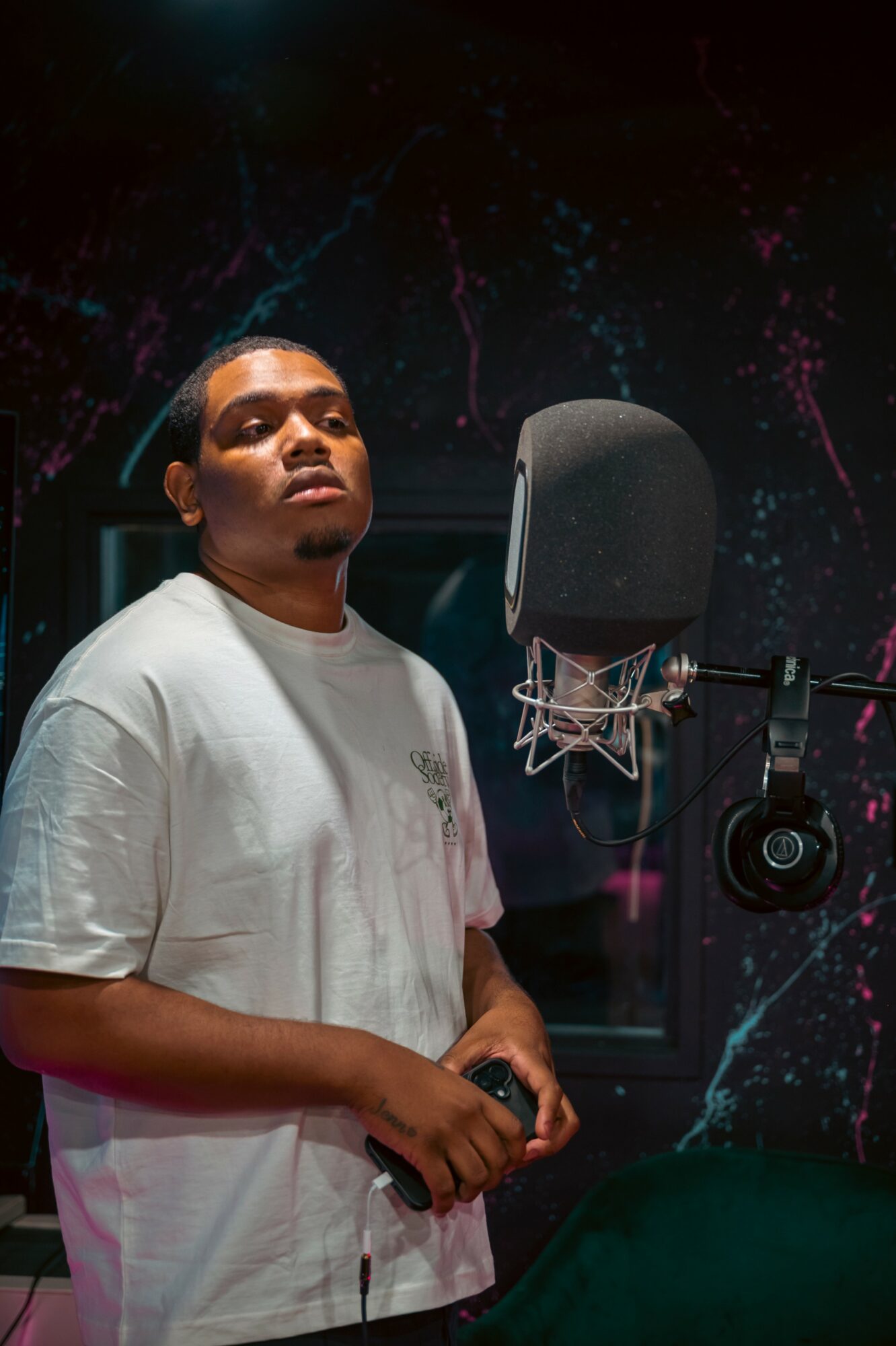 Young man in a recording studio with a microphone, dark background with colorful splashes, holding a phone, looking at camera.