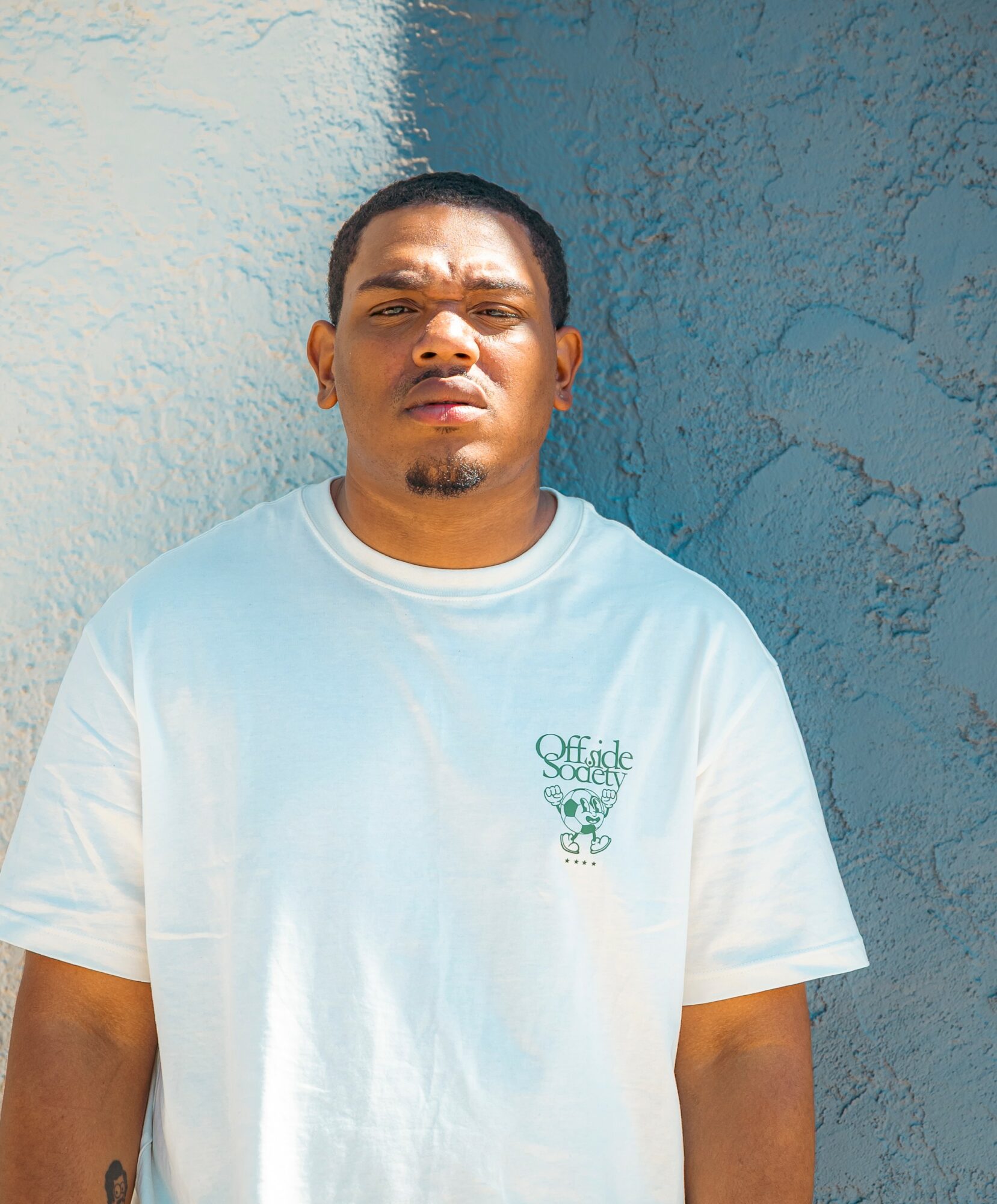 Young man with short hair and a goatee, wearing a white T-shirt, standing outdoors against a textured wall.