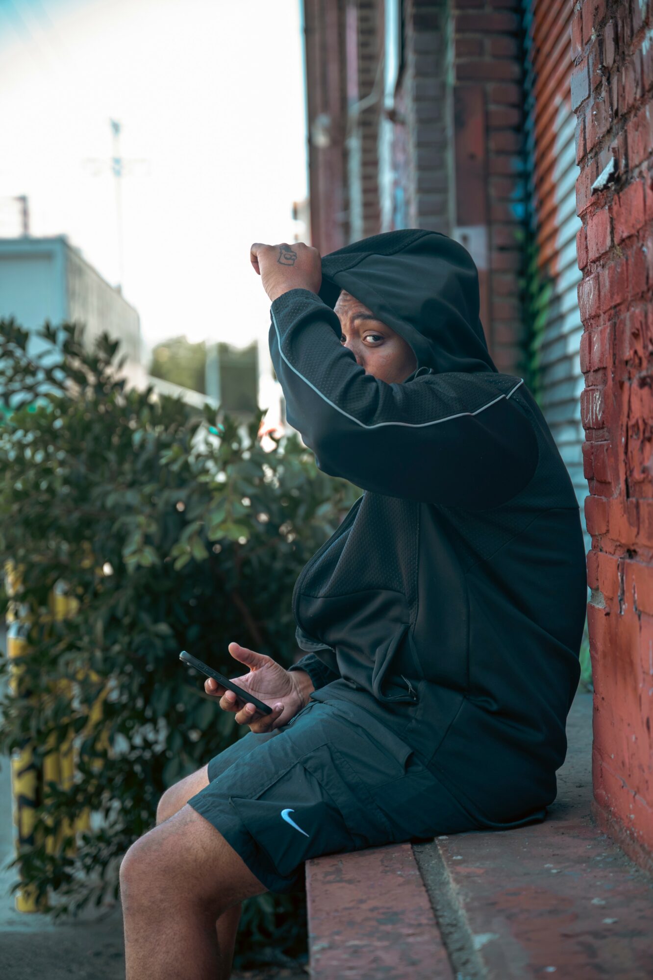 Child sitting on a ledge, holding a phone, wearing a hoodie, with a brick wall and greenery in background.
