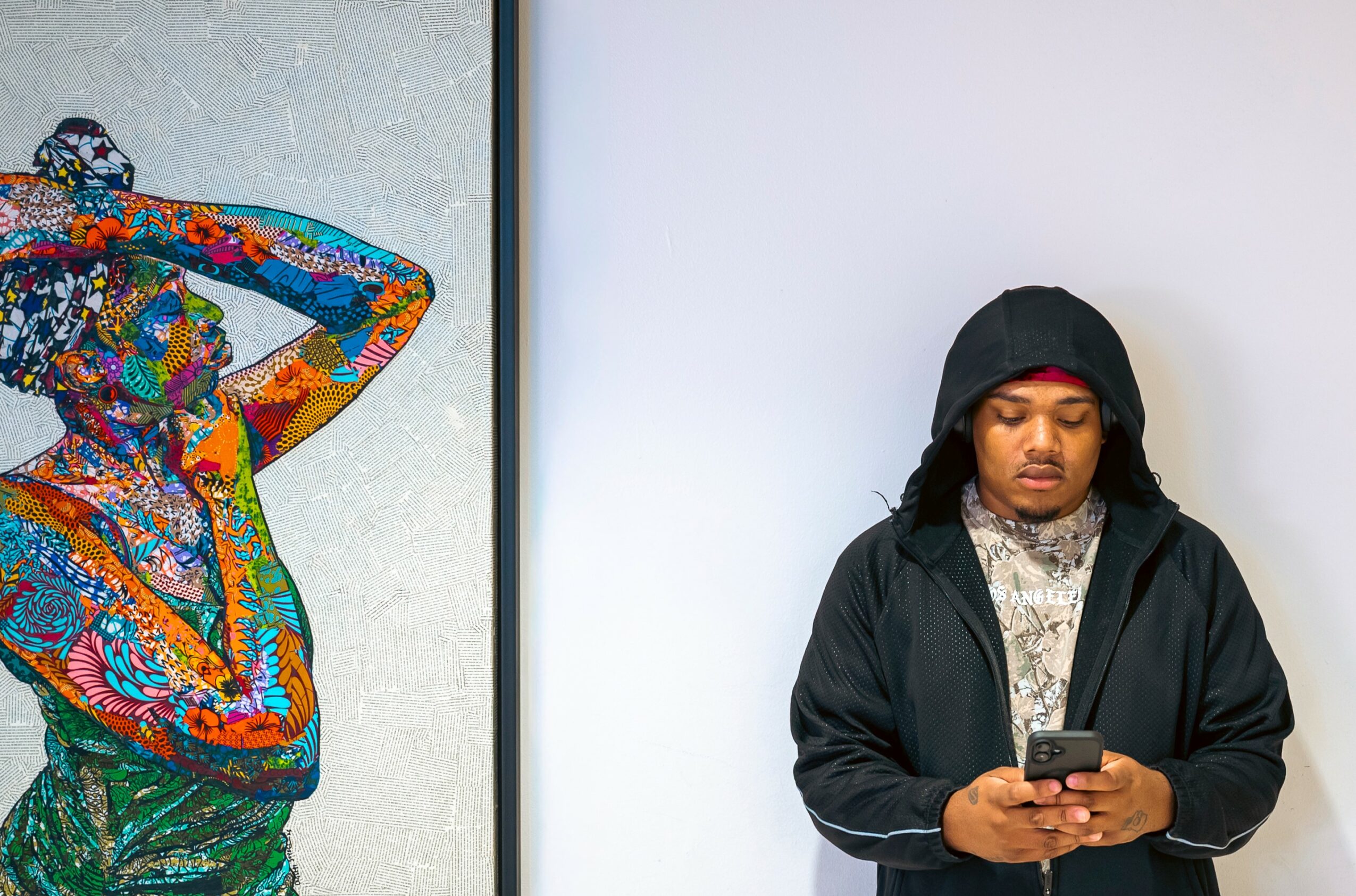 Person with long dark hair wearing a black hoodie looking at a phone, standing next to colorful artwork, against a plain background.