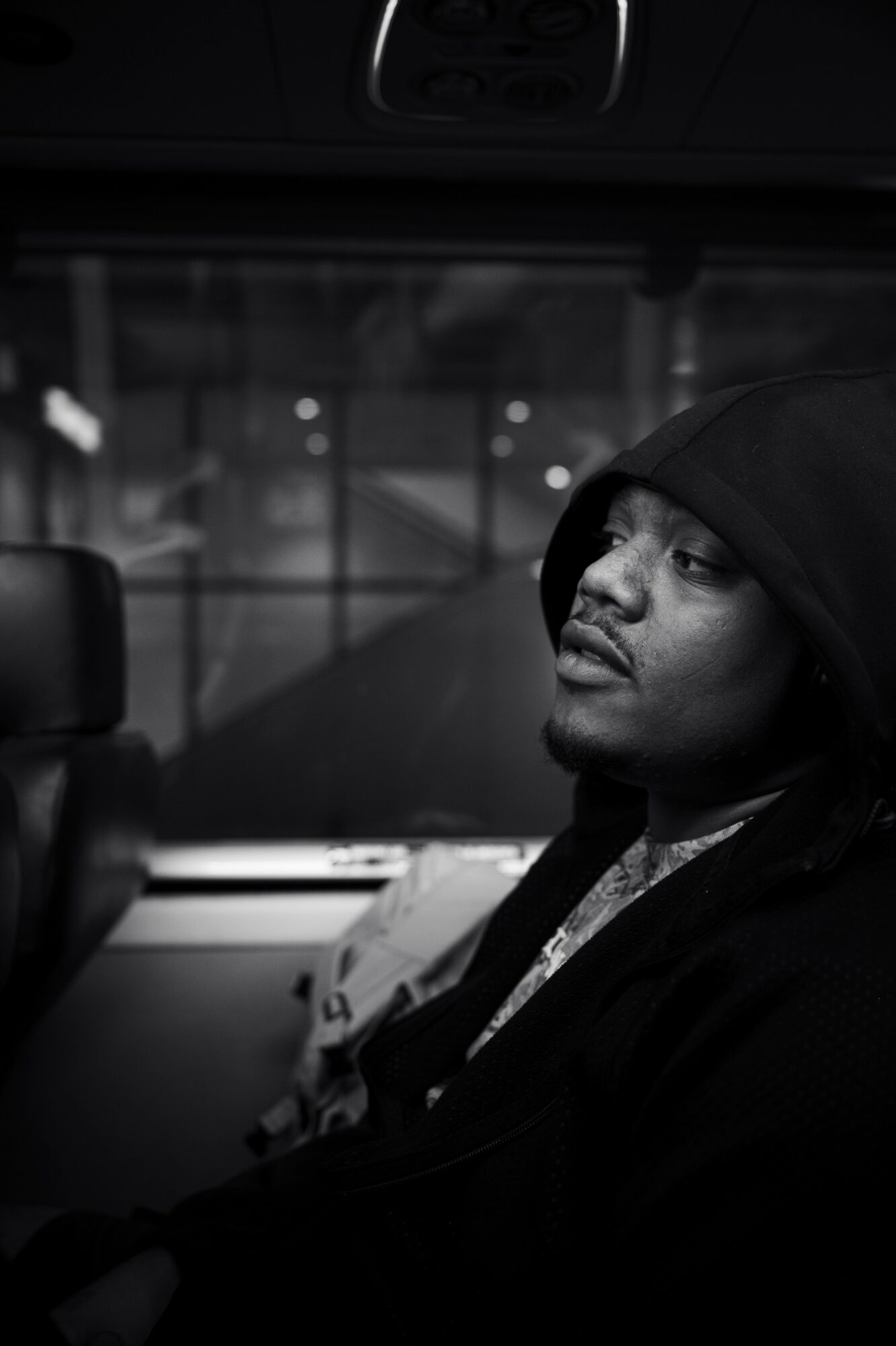 Man wearing a hoodie sitting in a vehicle, looking to the side, with a blurred background outside the window.