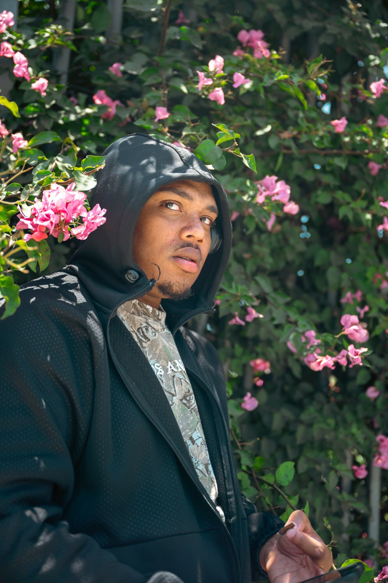 Person wearing a black hoodie and jacket, standing among pink flowering bushes outdoors, looking to the side.