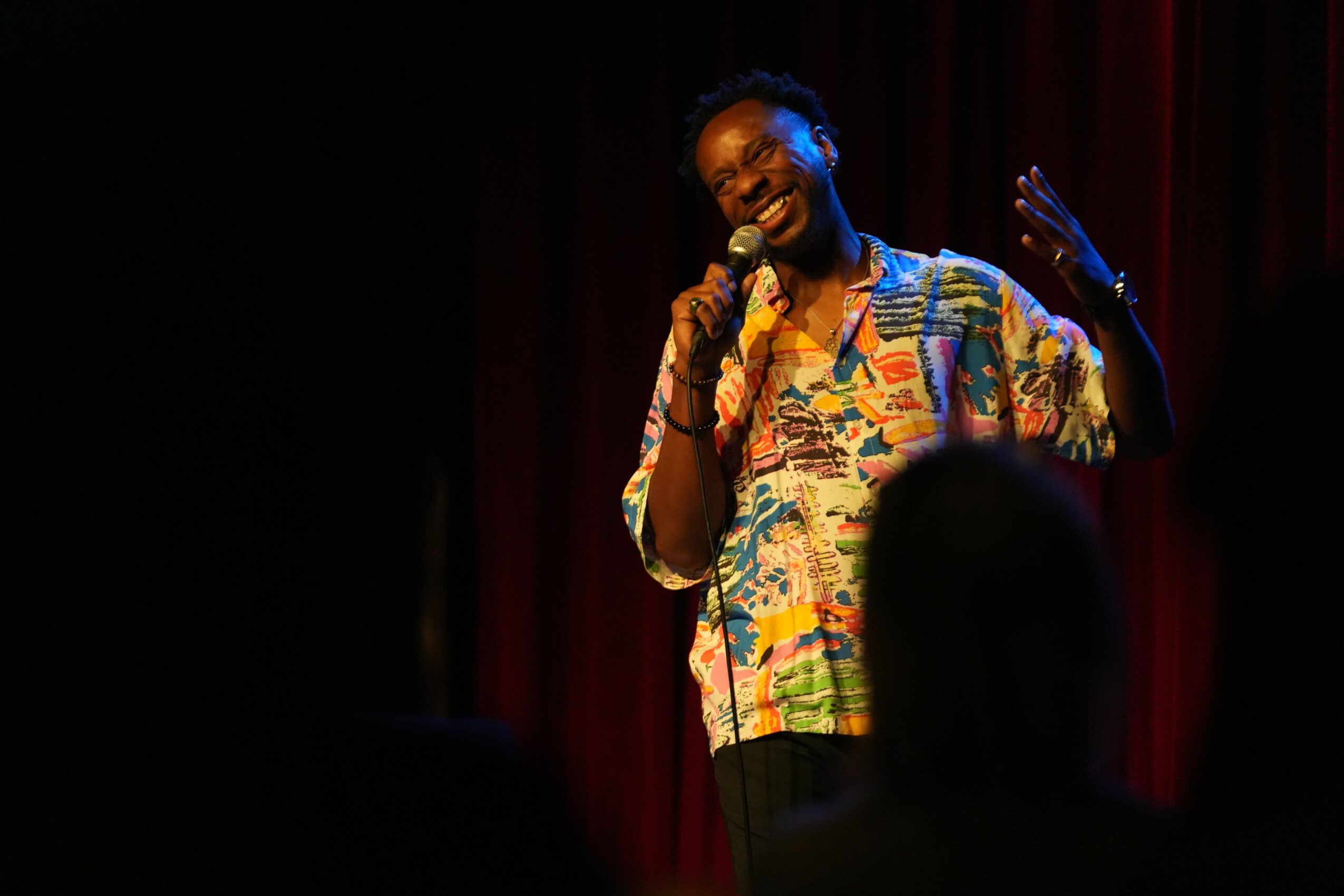 Person with braided hair singing into a microphone on stage, wearing a colorful patterned shirt, with a red curtain background.