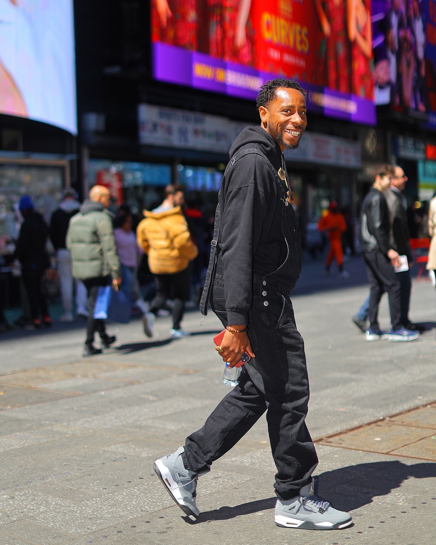Smiling man walking on busy city street with people and digital billboards in background.