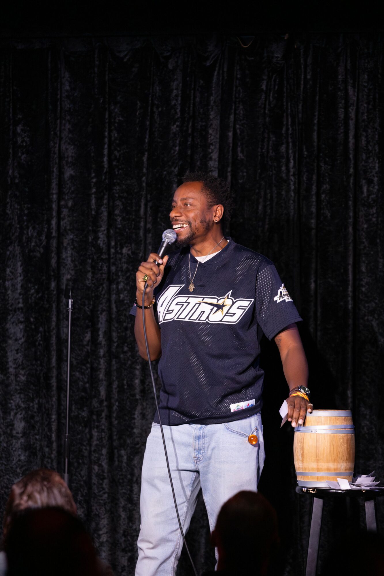 Man holding microphone on stage with black curtain background, smiling, wearing a dark sports jersey and light jeans.