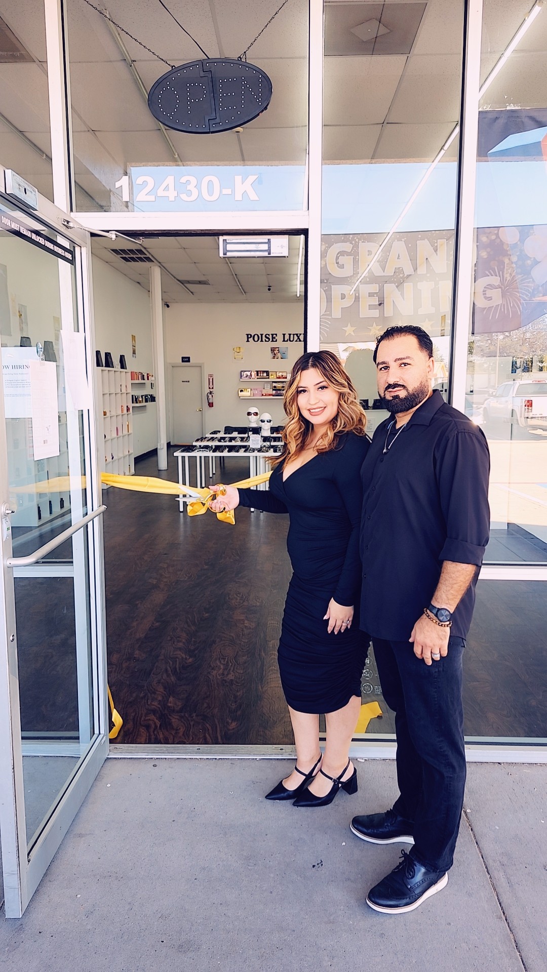 Two people stand outside a store, smiling, with a glass door and store interior visible behind them.