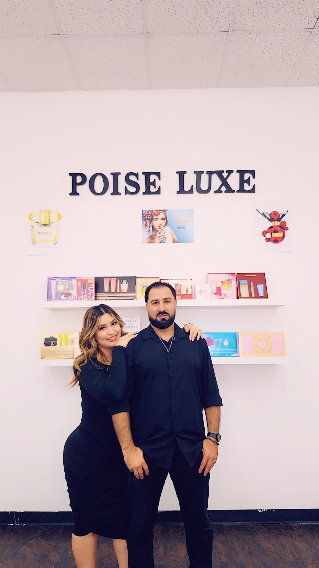 Two people stand in front of a white wall with shelves and decorations, smiling at the camera.