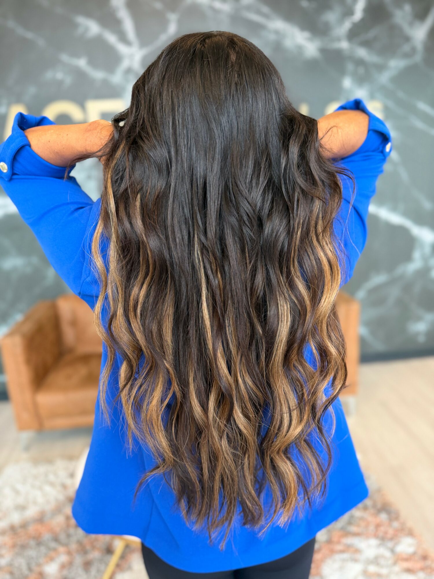 Person with long, wavy hair wearing a blue top, facing away, with hands behind head, in front of a textured wall.