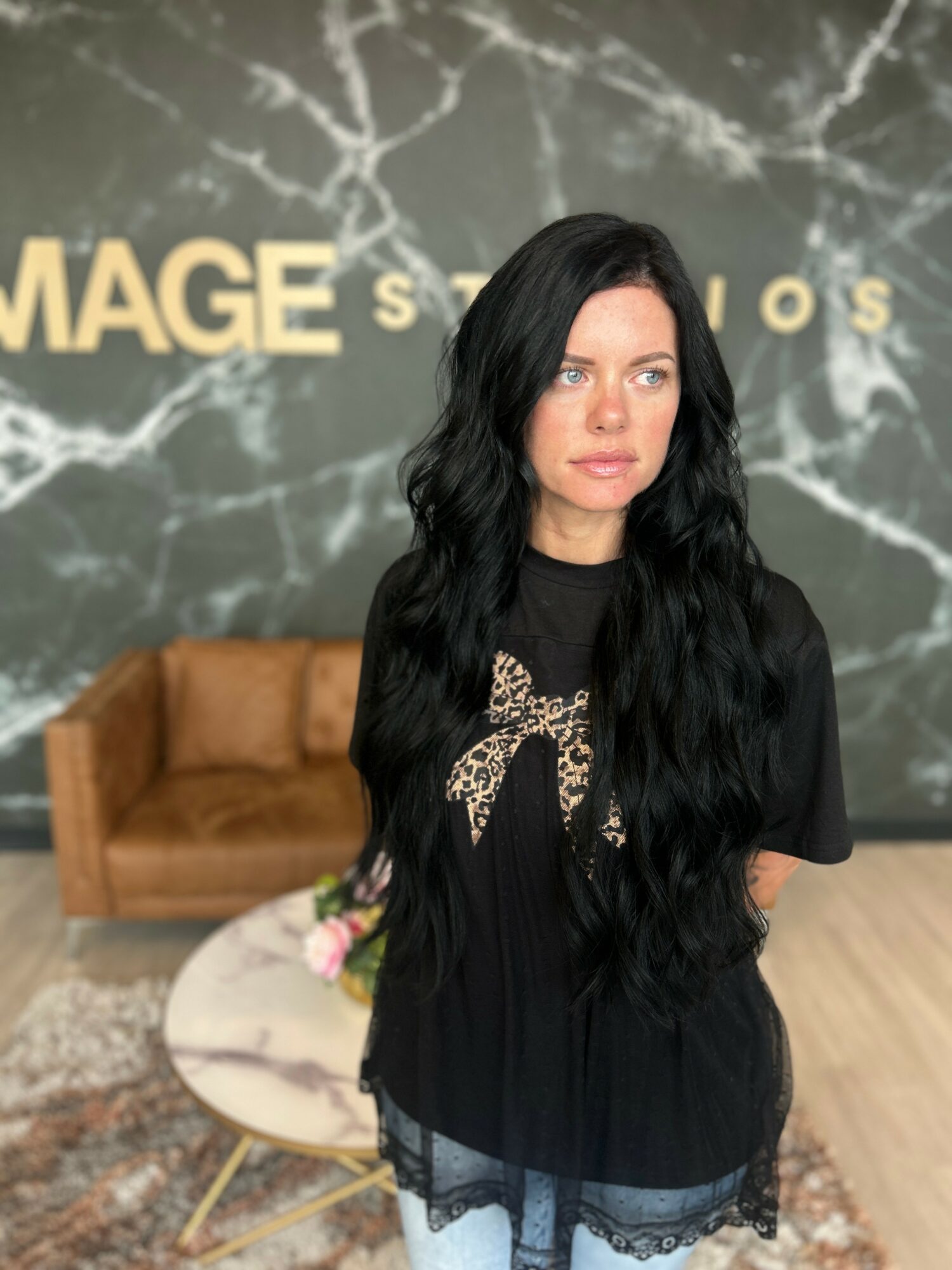 Woman with long black hair and leopard print bow on black top, standing indoors with marble wall background and brown sofa.