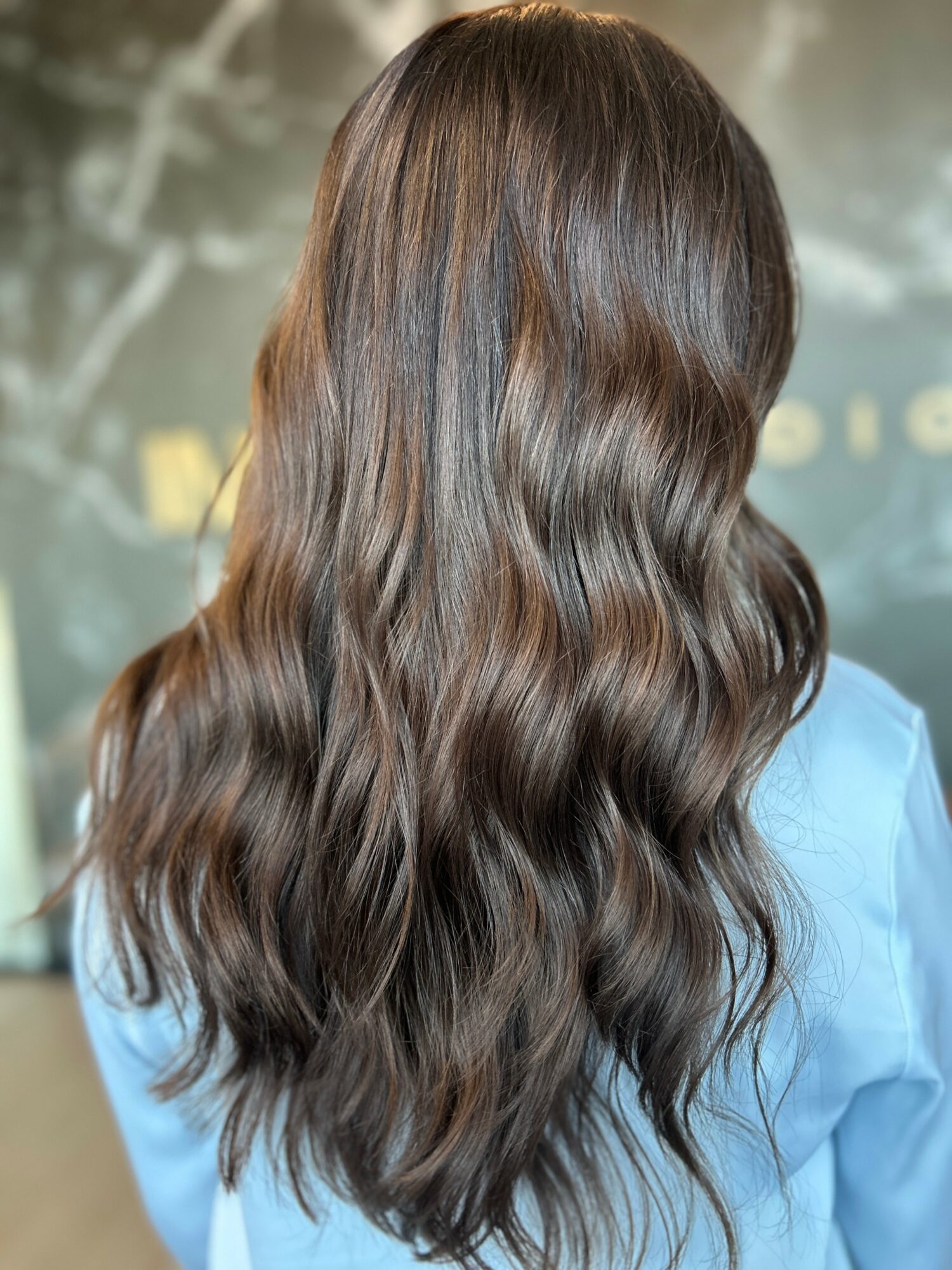 Back of a person with long, wavy brown hair, wearing a light blue shirt, in a blurred background.