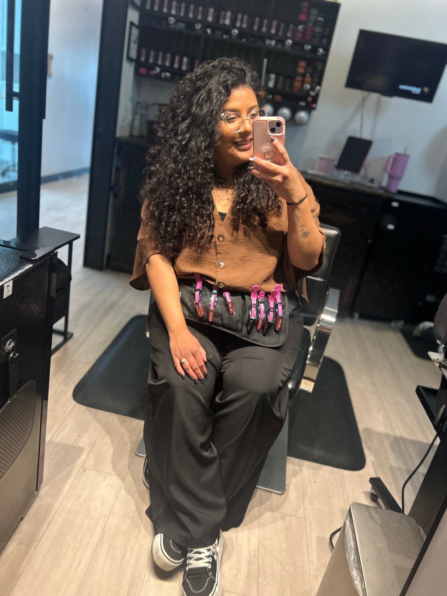 Woman with curly hair taking a mirror selfie in a salon, wearing a brown top and black pants, holding a phone.
