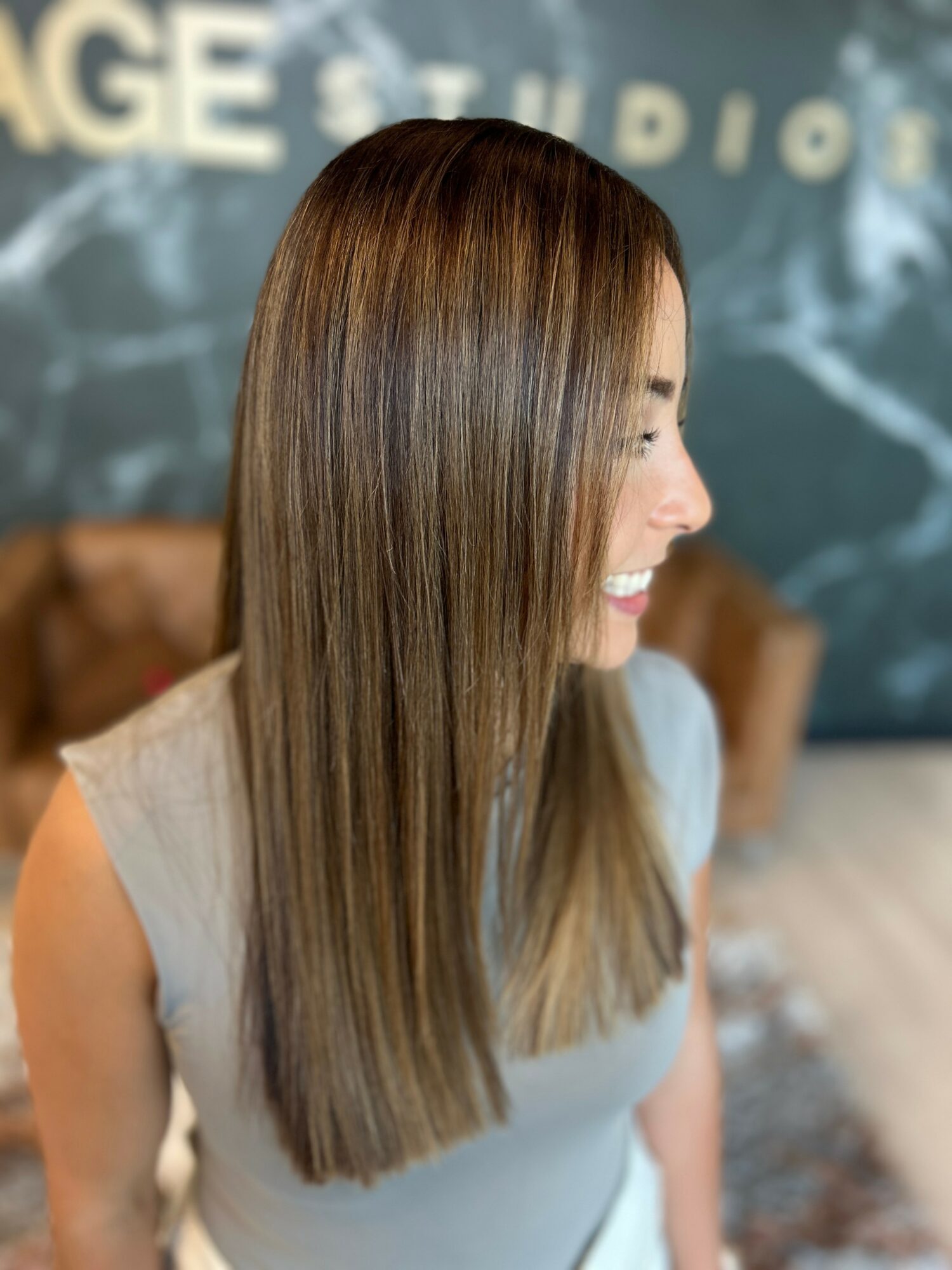 Side view of a woman with long, straight, brown hair smiling, wearing a light-colored top, in an indoor setting.