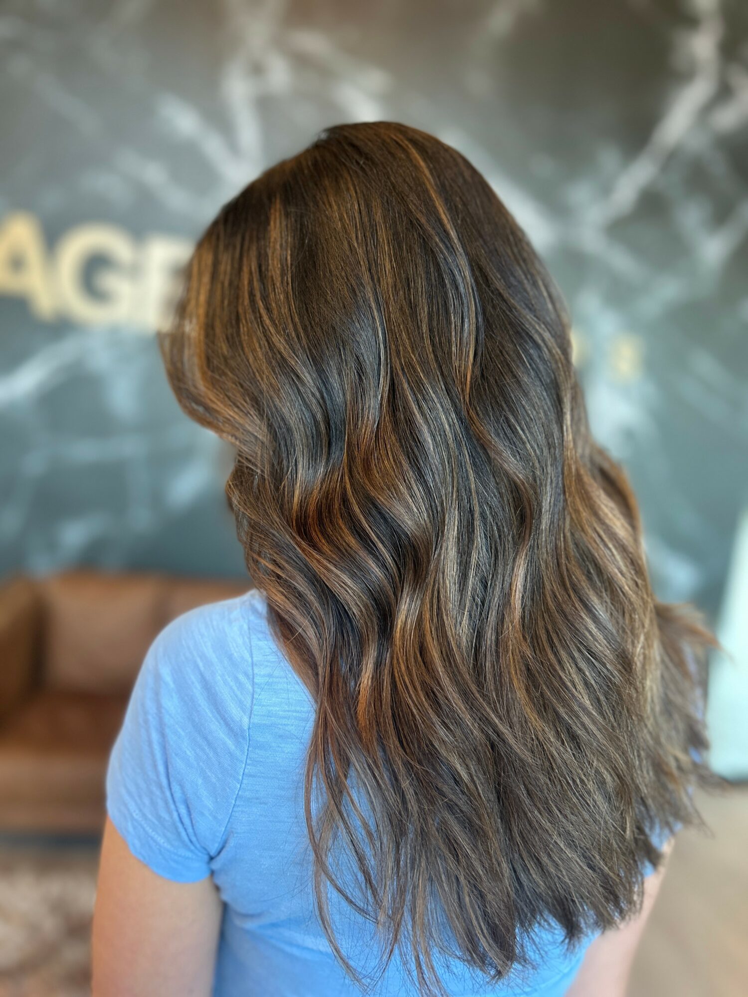 Back view of a woman with long, wavy brown hair wearing a blue shirt, standing indoors with a blurred background.