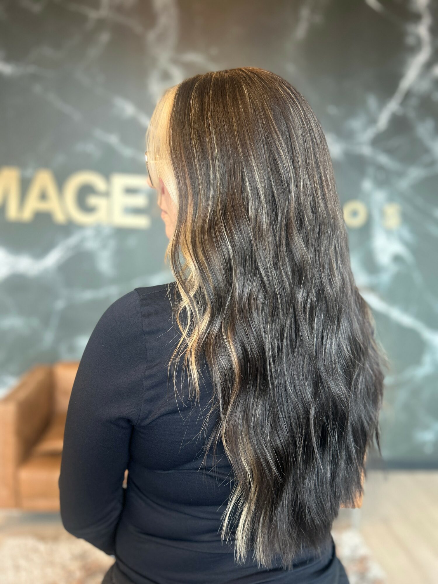 Back view of a woman with long, wavy hair standing indoors, with a blurred background and partial text visible.