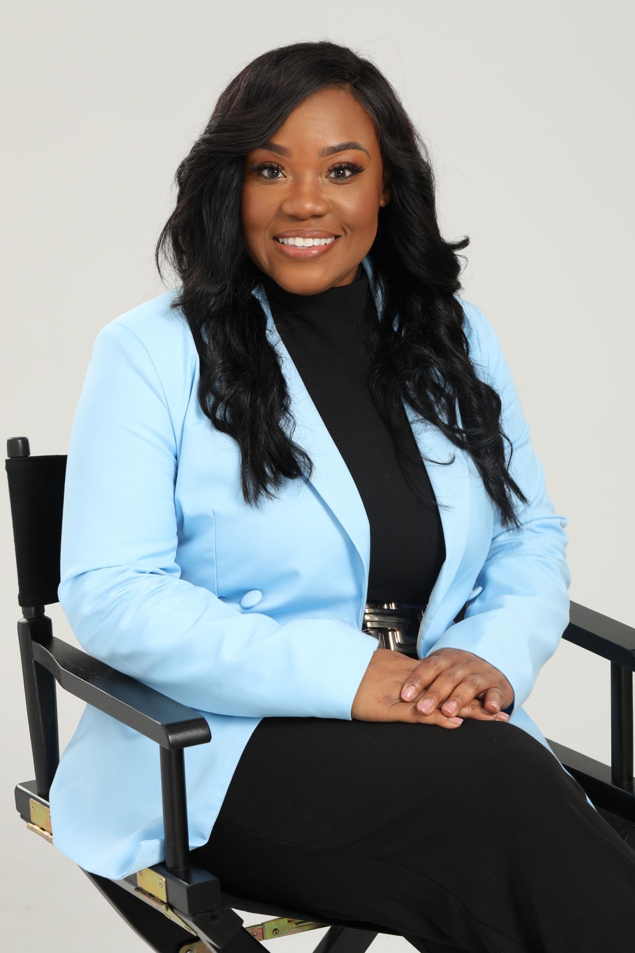 Woman sitting on a chair, smiling, wearing a light blue blazer and black pants, with long black hair.
