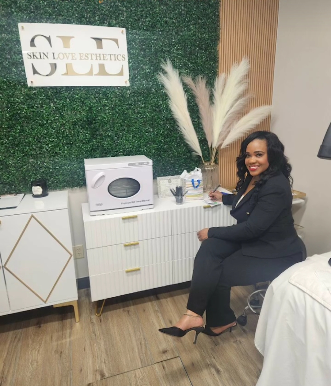Woman in black suit sitting at a desk with a white cabinet, green wall, and decorative plants behind her.