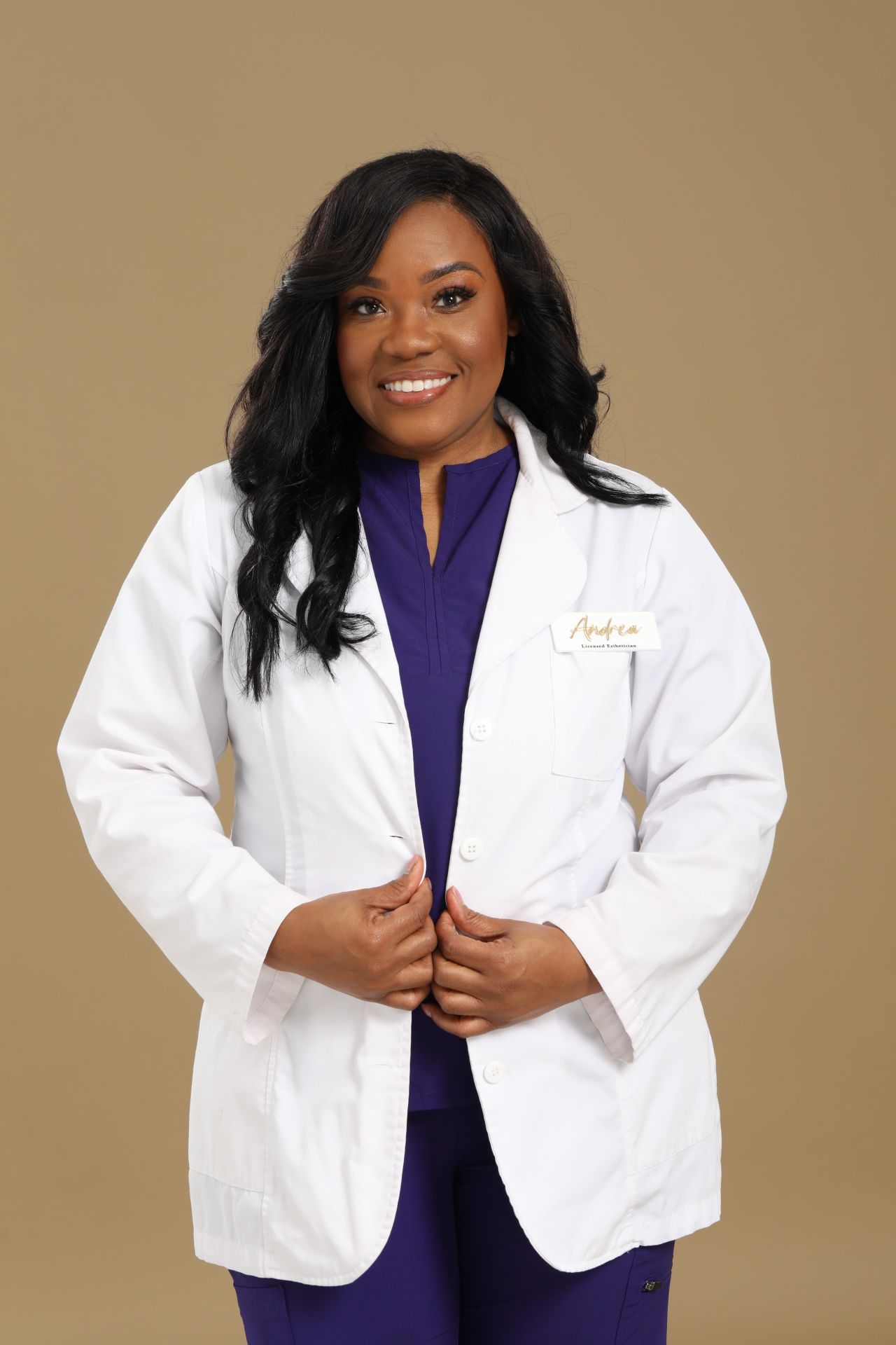 Woman in white coat smiling, standing against a beige background, with long black hair and dark eyes.