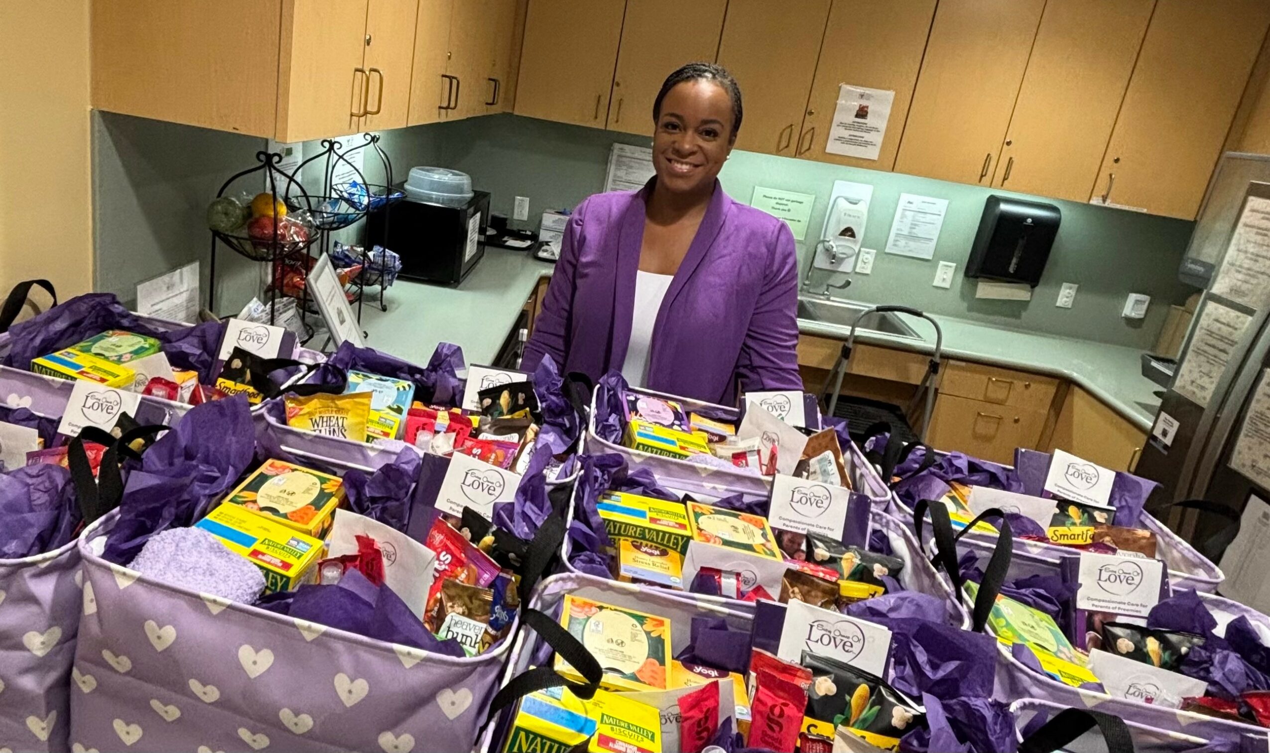 Smiling woman in purple blazer standing behind tables filled with gift bags and boxes in a room with cabinets.