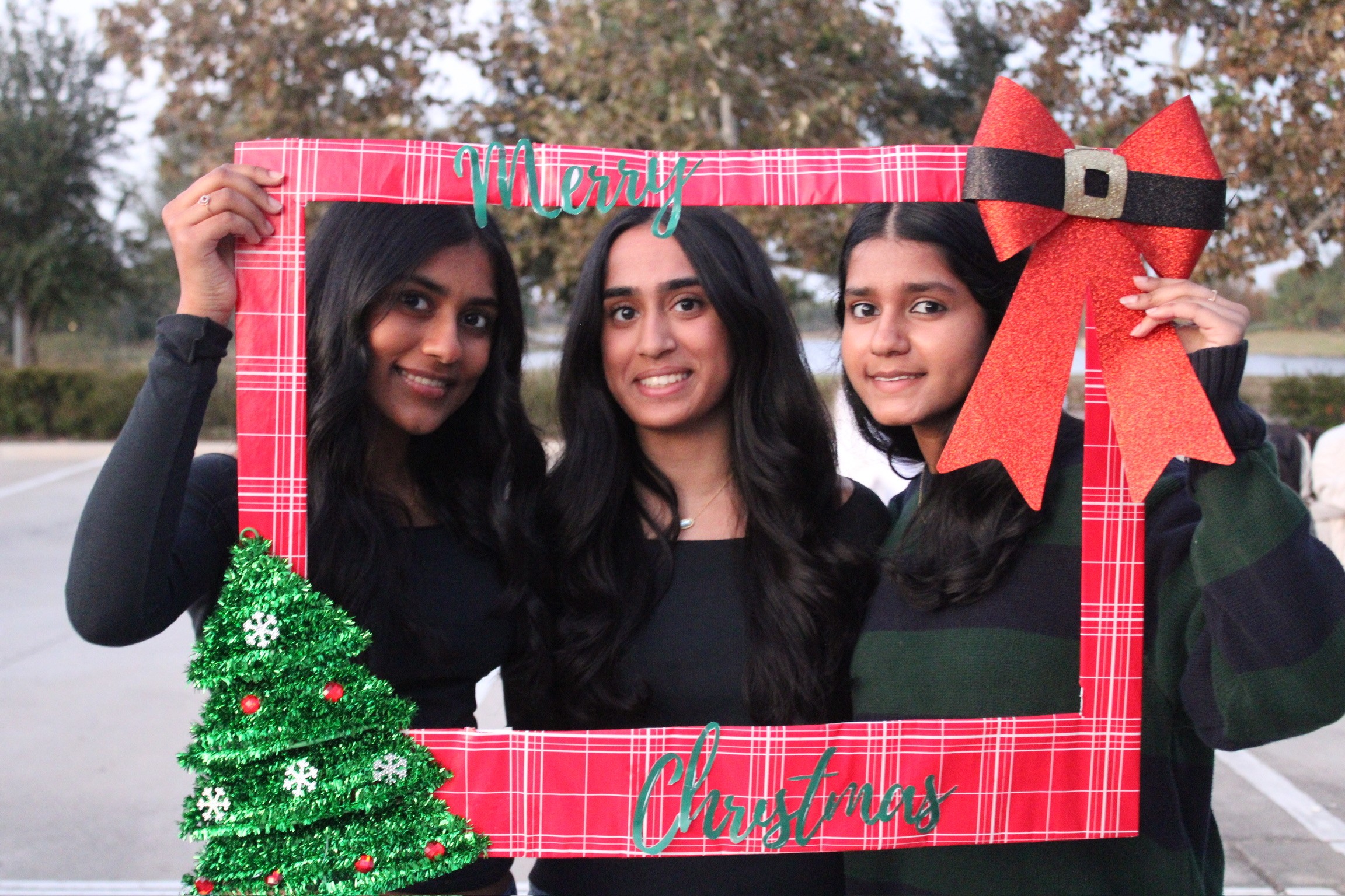 Three women holding a Christmas-themed photo frame outdoors, smiling, with a Christmas tree decoration on the frame, and a large red bow.