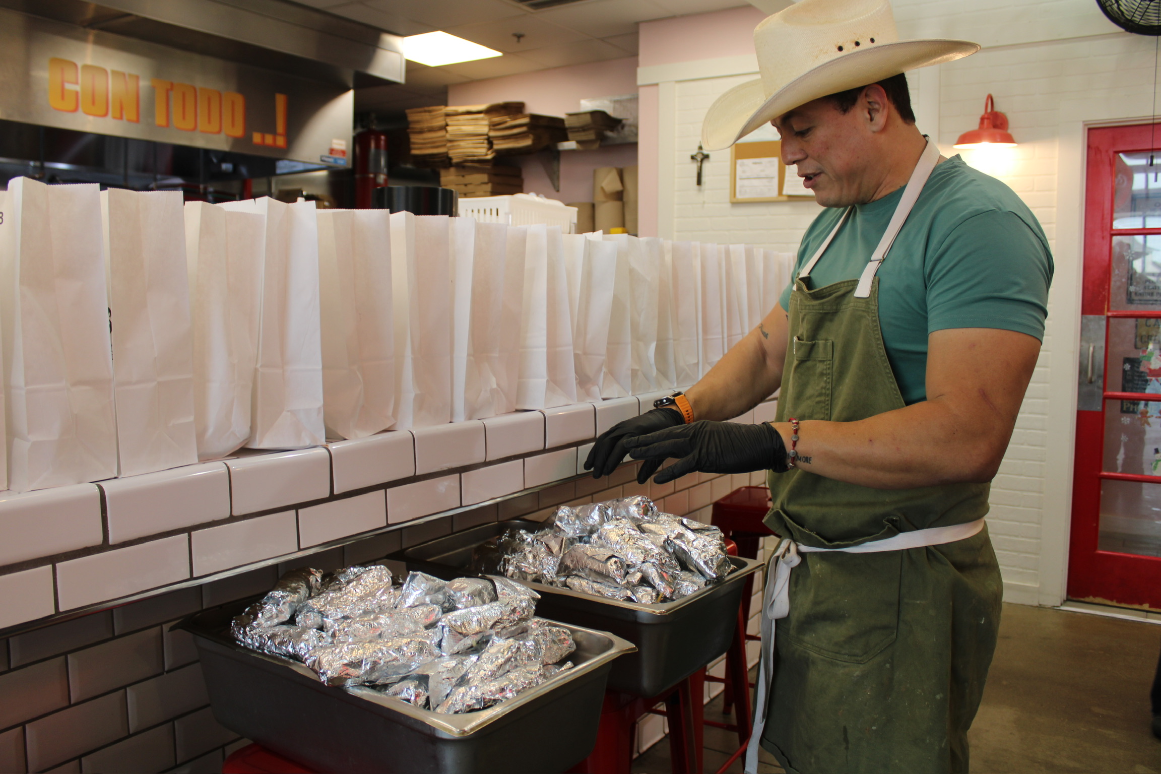 Man wearing a cowboy hat and apron prepares food in a restaurant kitchen, with foil-wrapped items on the counter.