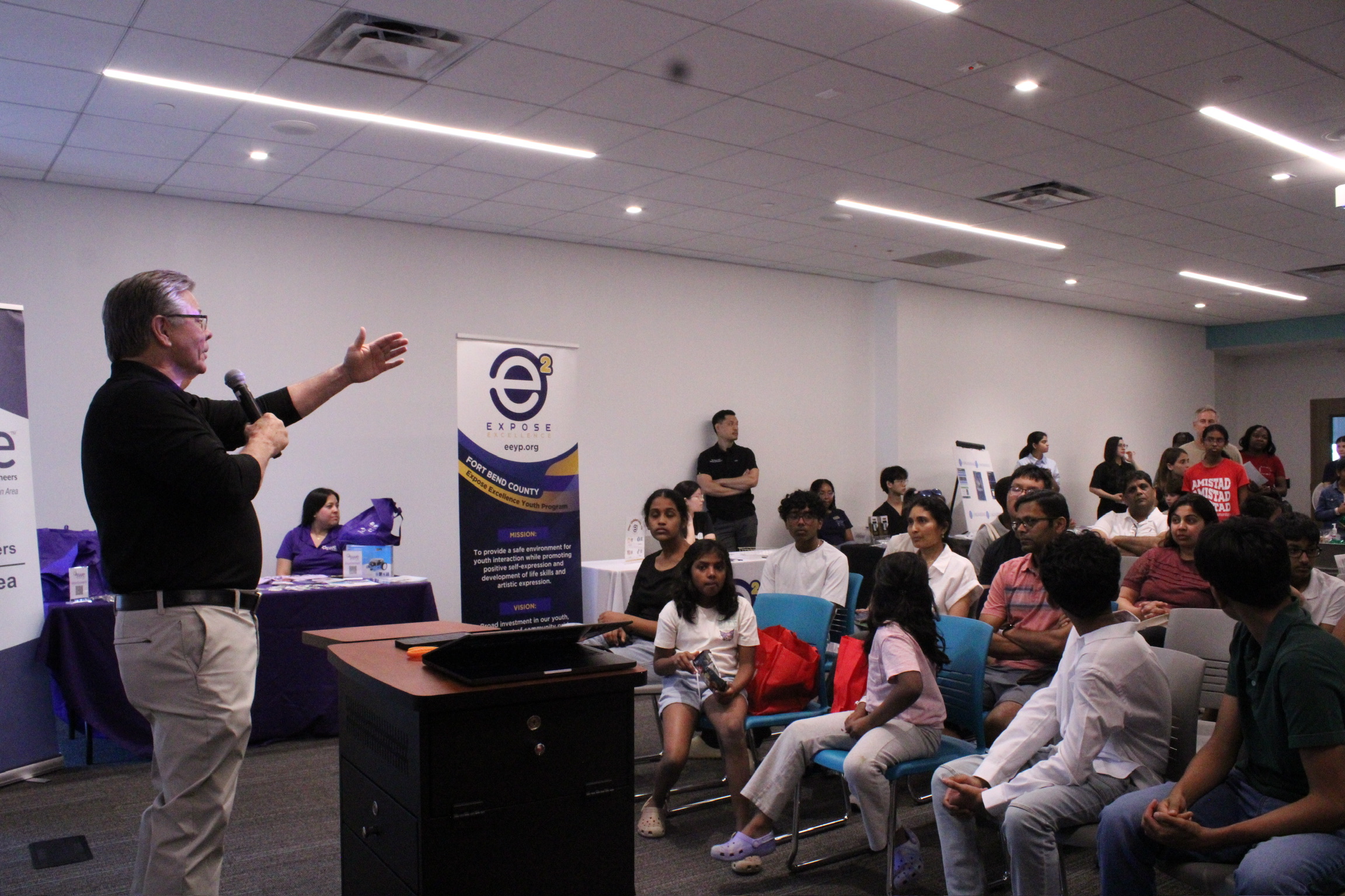 A man is speaking to a group of people seated in a room with banners and tables, some people are taking notes.