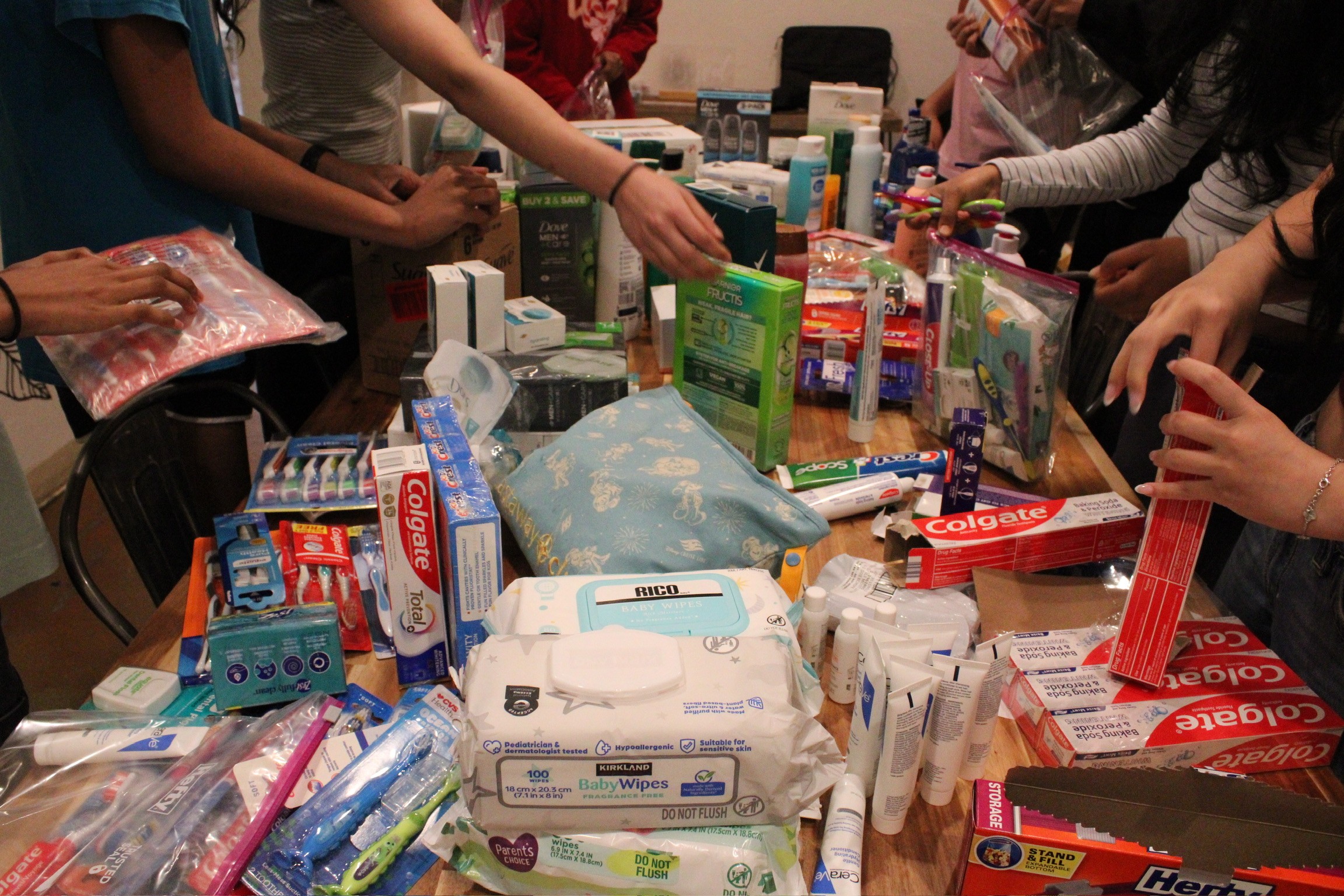 People gather around a table filled with various household and personal care products, reaching for items.