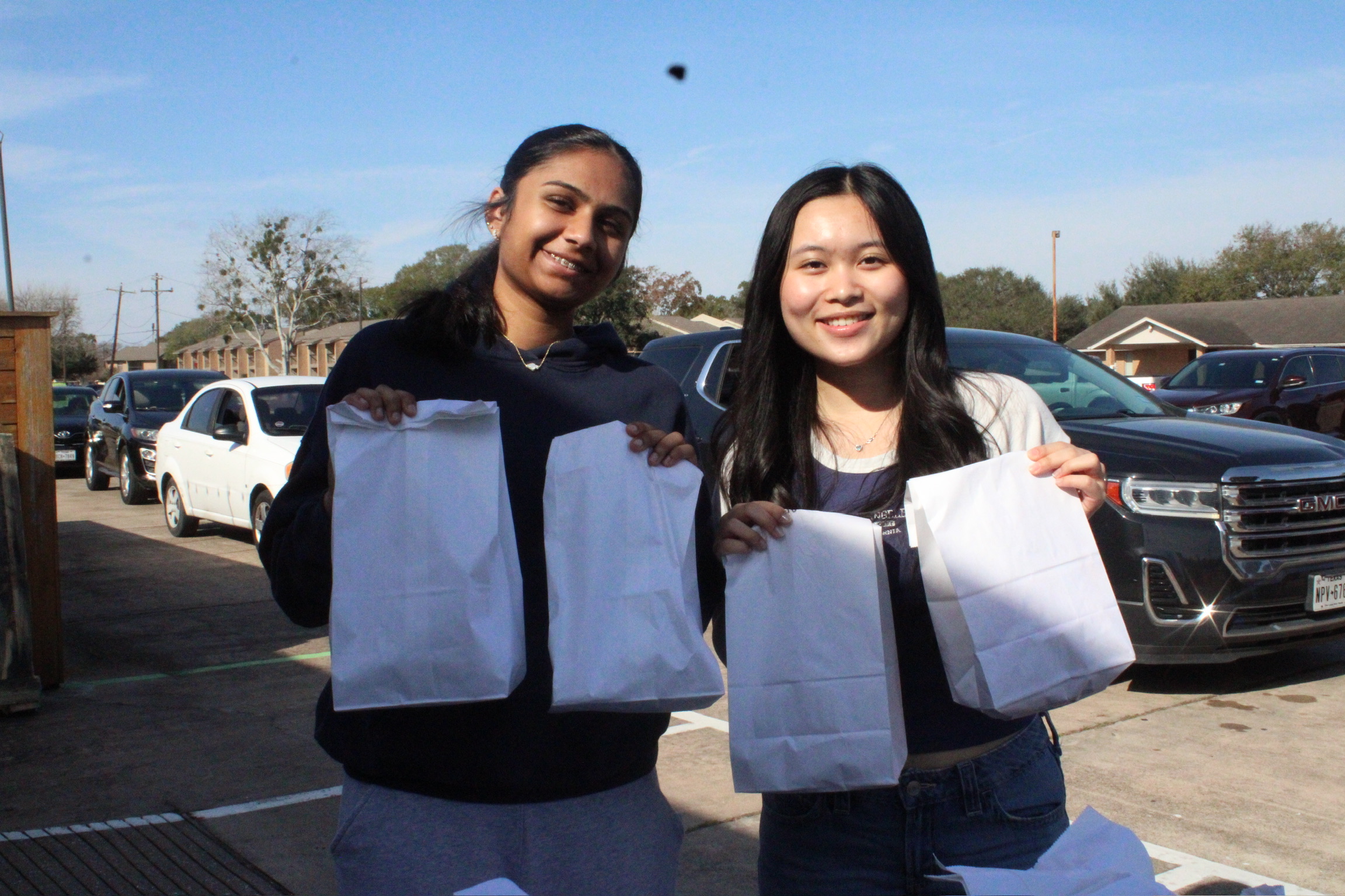 Two smiling women holding paper bags outdoors in a parking lot, with cars and buildings in the background.