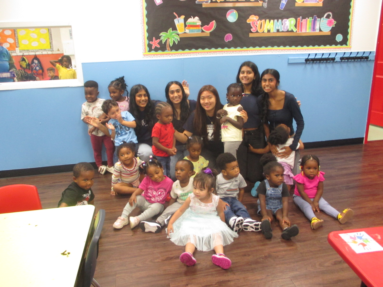 Group of young children and adults in a classroom, some sitting on the floor and others standing, with colorful decorations on the wall.