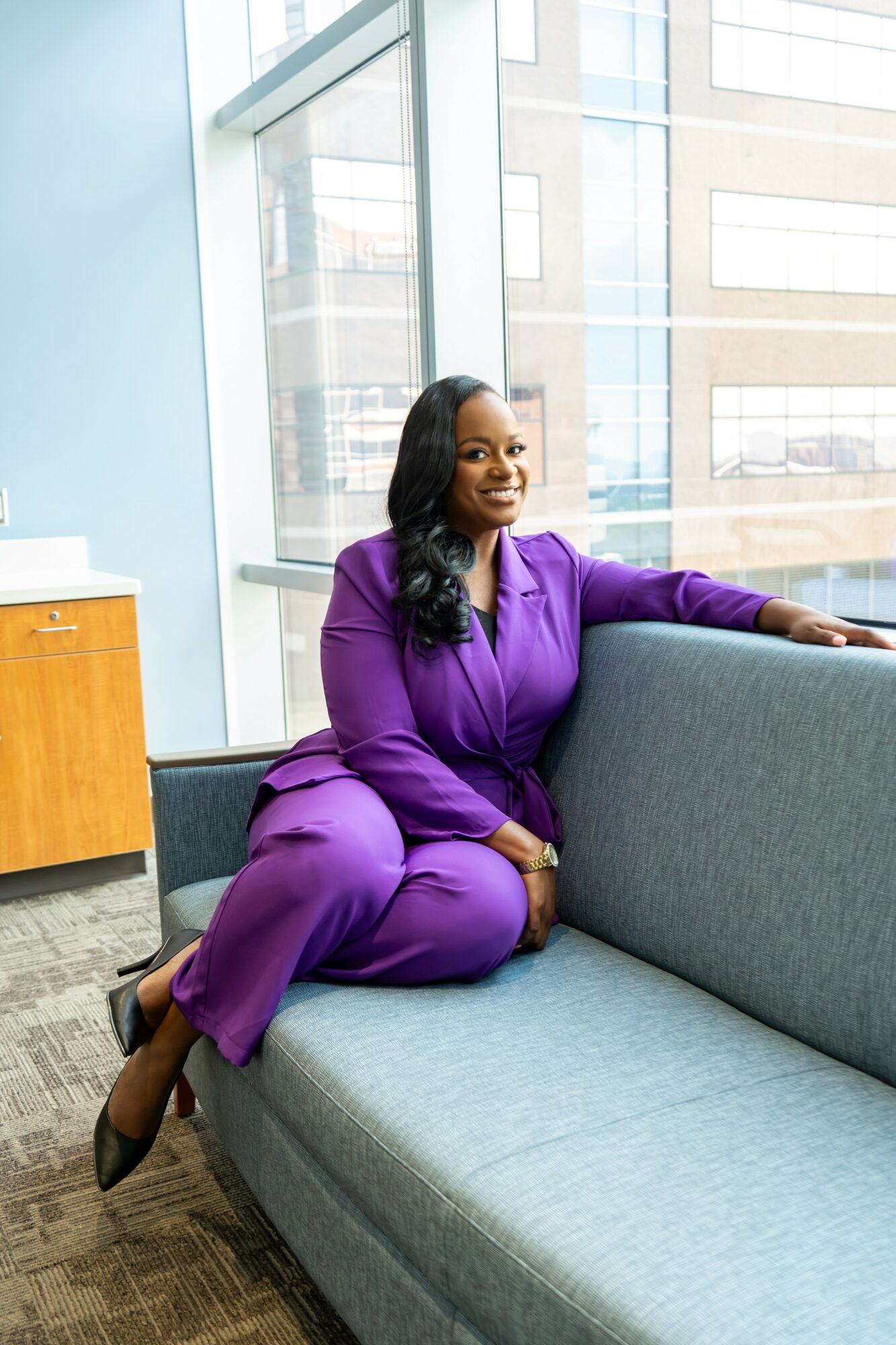 Woman in purple suit sitting on a gray sofa near large windows, smiling, with a wooden cabinet nearby.