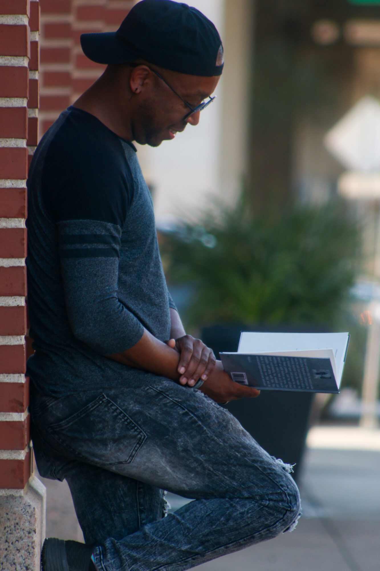 Person leaning against a brick wall using a laptop outdoors, wearing a cap and glasses, with blurred background.