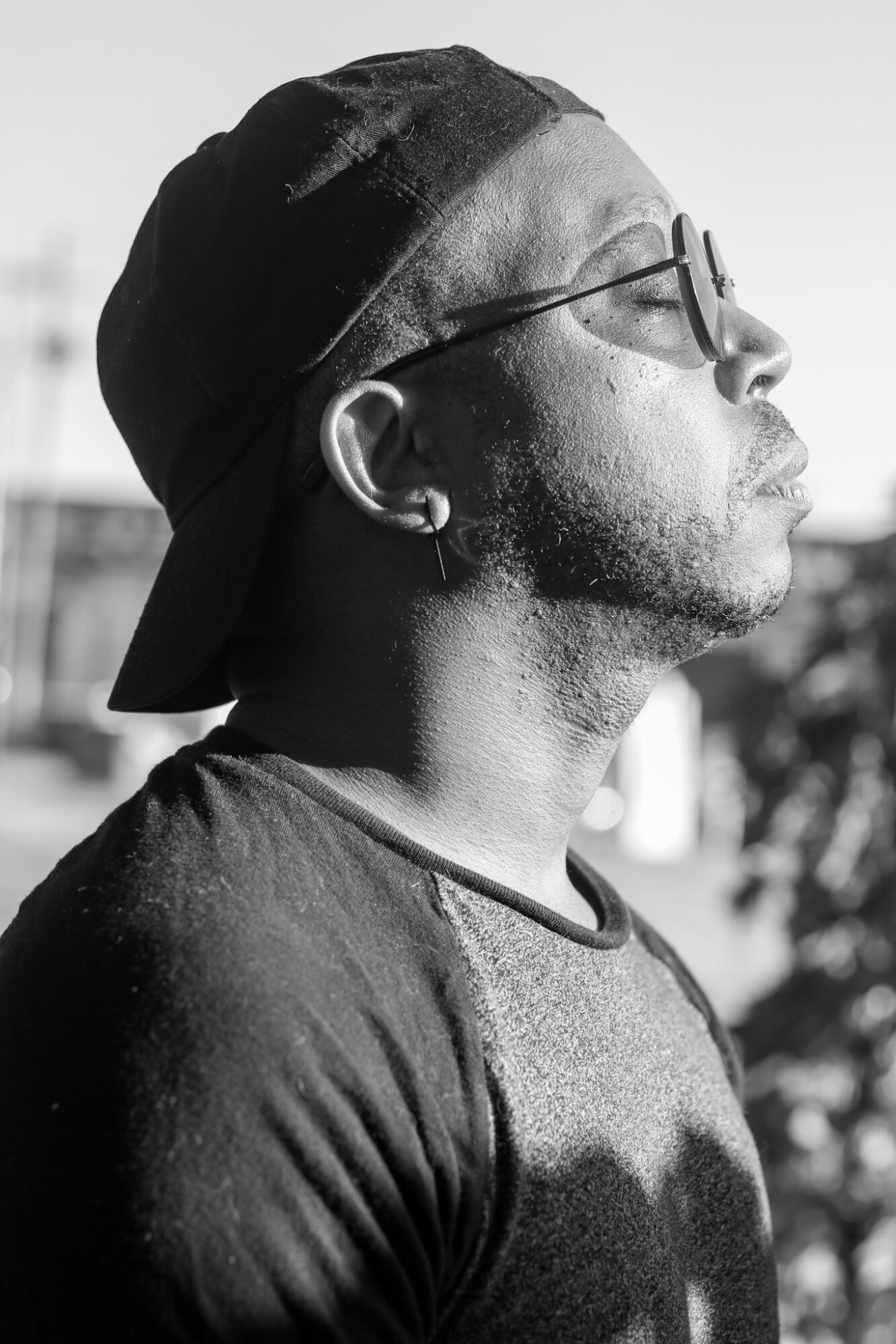 Side profile of a man wearing sunglasses, a cap backward, and a T-shirt outdoors, with sunlight and shadows.