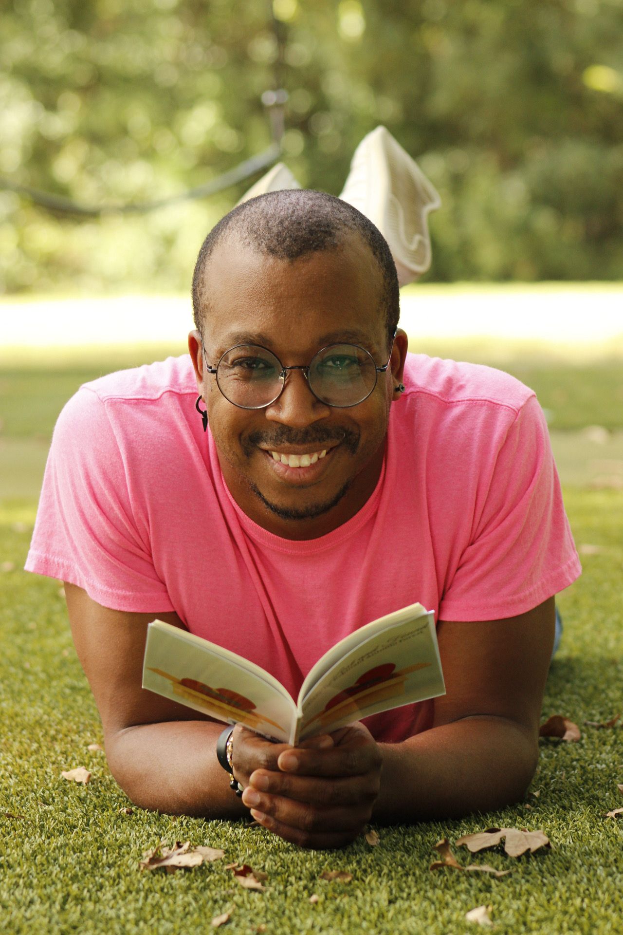 Person lying on grass holding a book, smiling, wearing glasses and a pink shirt, outdoors with trees in background.