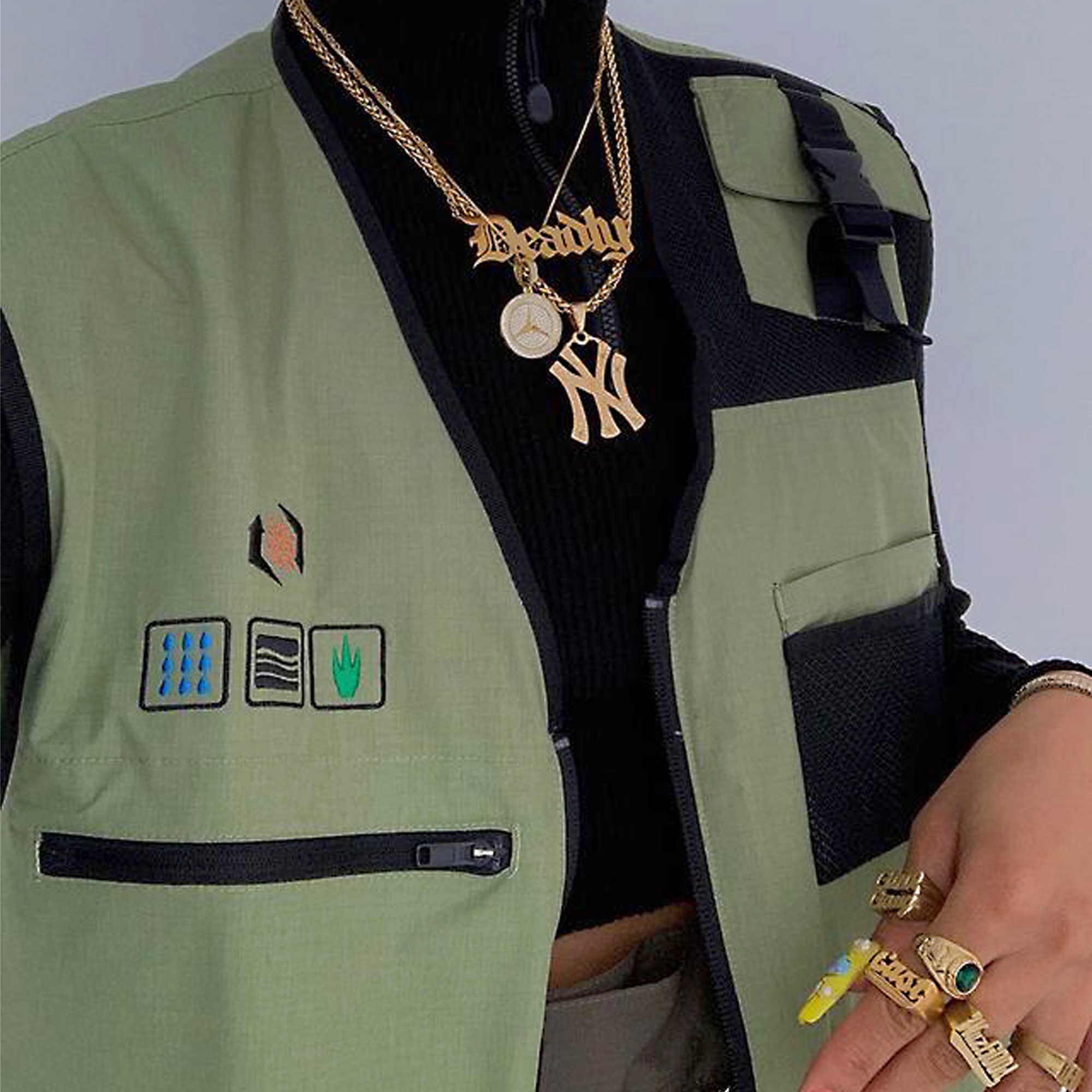 Close-up of a person wearing a green vest with patches, layered over a black top, with gold jewelry and rings, against a plain background.