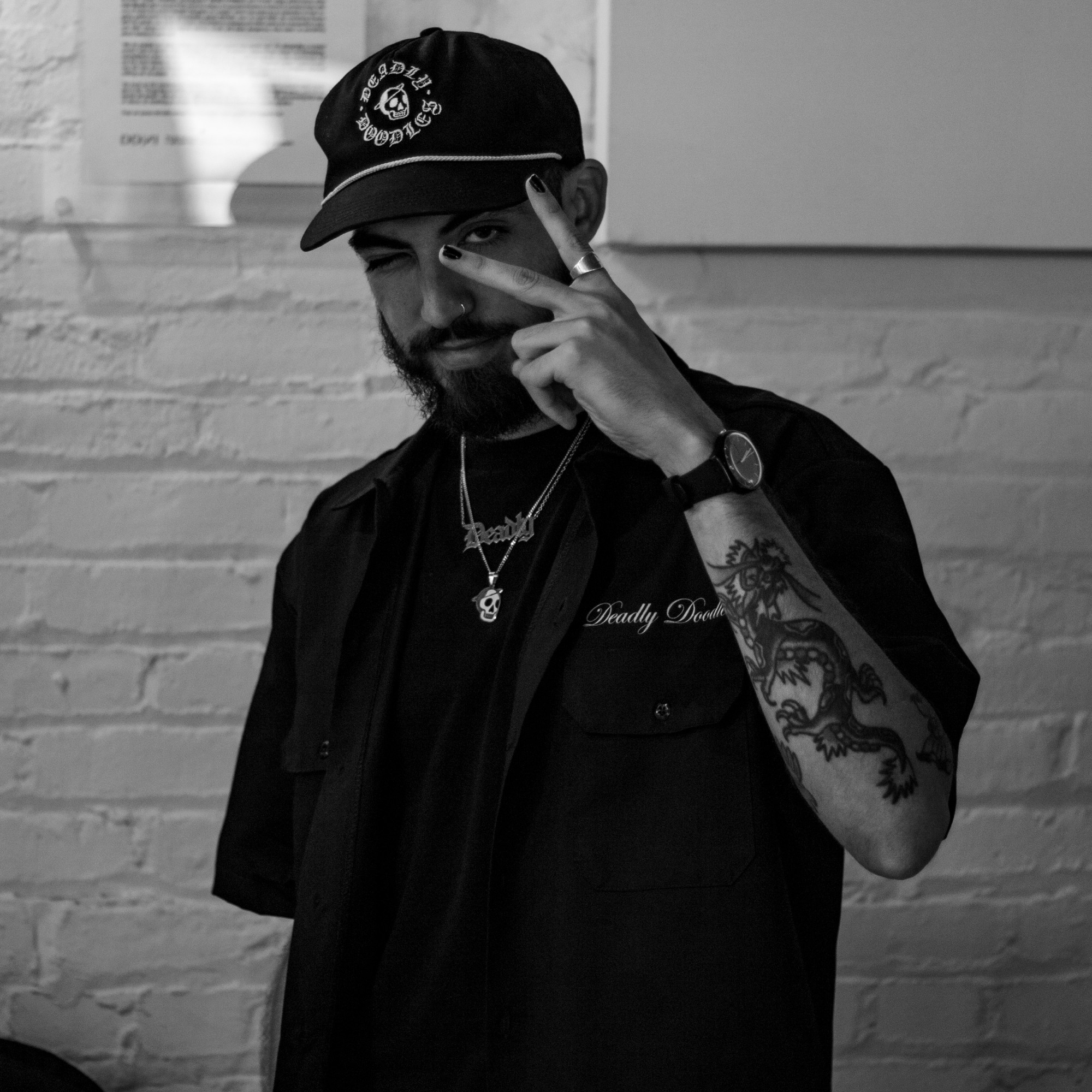 Man with a beard wearing a cap, pointing to his temple, standing against a brick wall, in black and white.