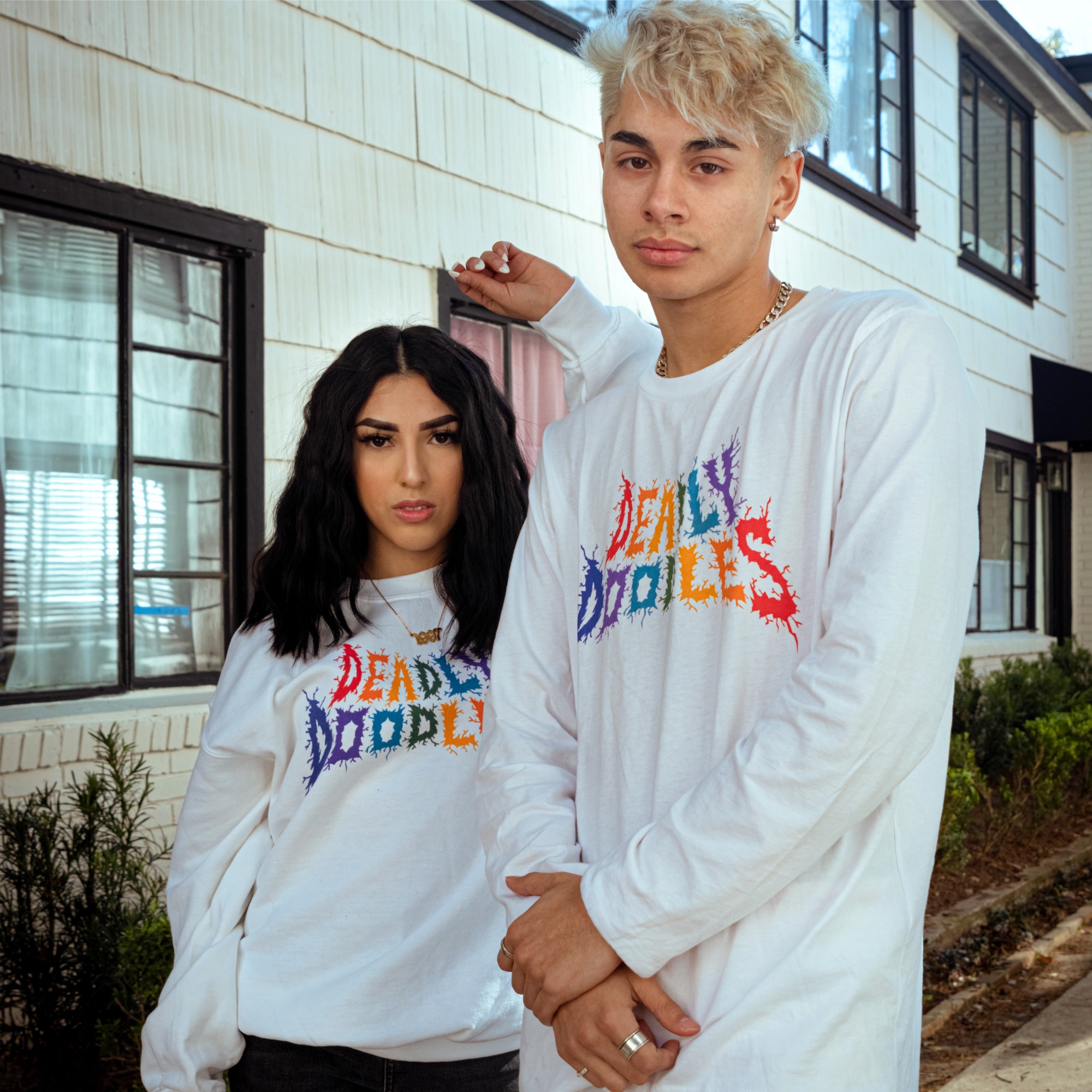 Two young people wearing matching white sweatshirts with colorful text, standing outside a building with large windows.