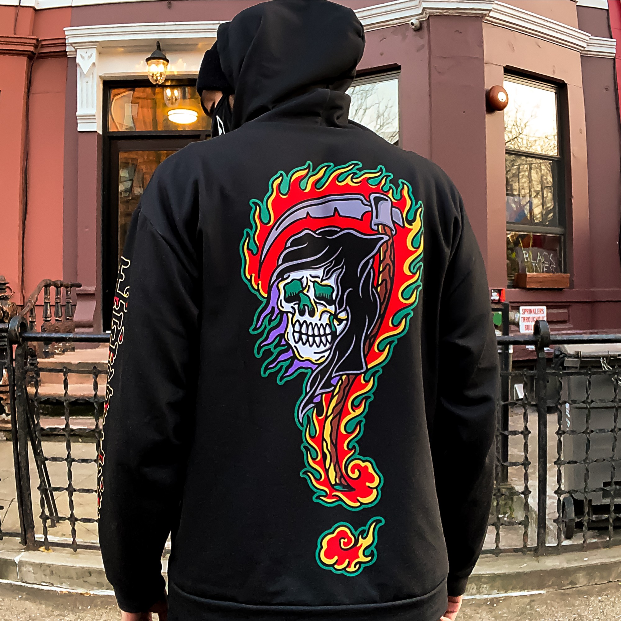 Person wearing a black hoodie with a colorful skull and question mark design on the back, standing outdoors near a building.